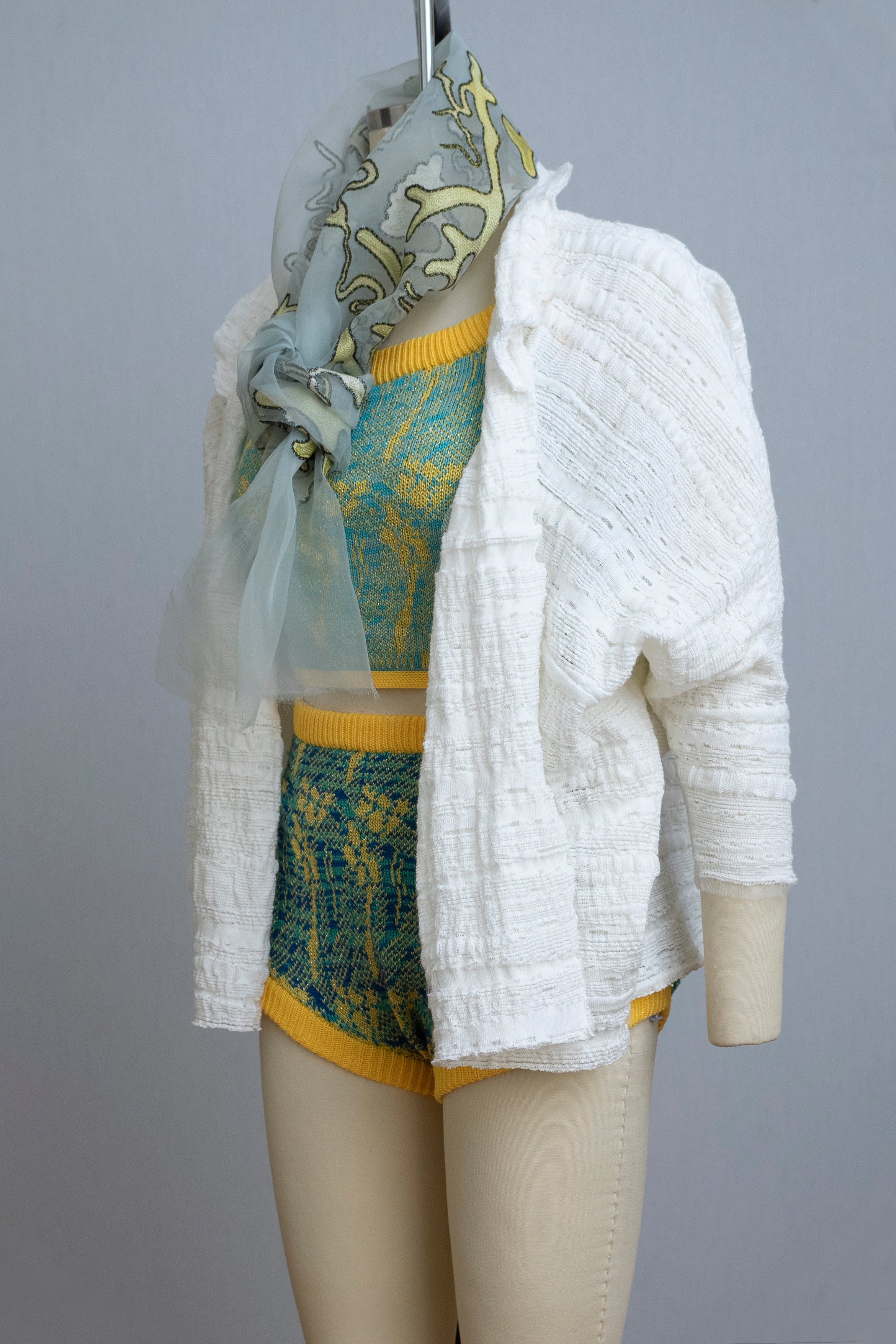 dress form wearing a knit playsuit in blues and yellows, a white knit sweater, and a sheer painted scarf