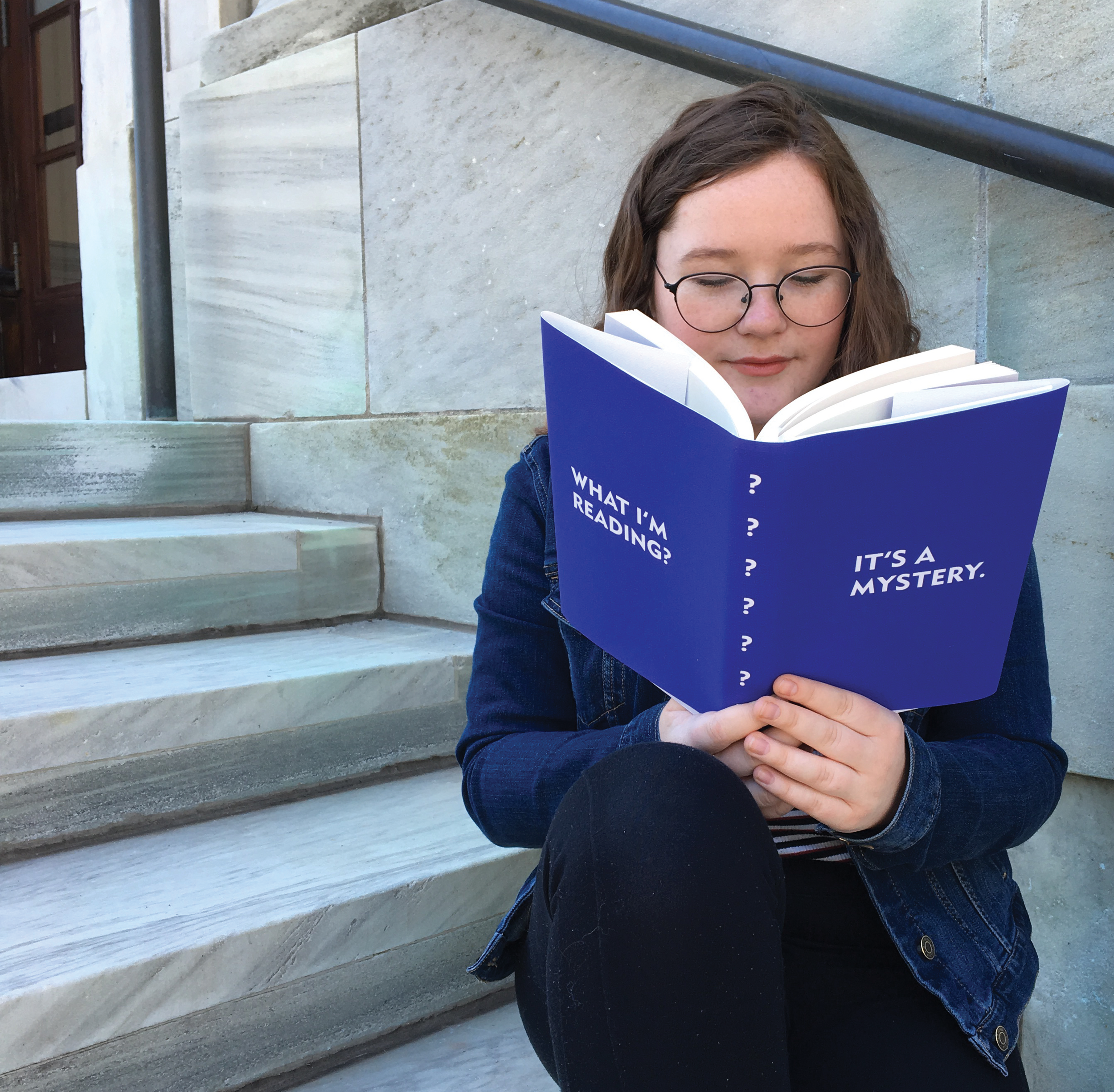 Woman reading a book with a blue cover with text 'What I'm Reading? It's a Mystery'