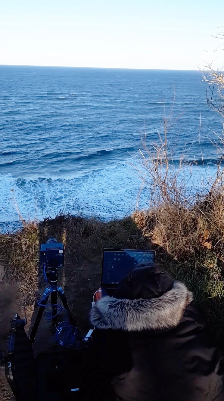 Person in heavy coat works with an Edgertronic Camera and laptop high above the sea.