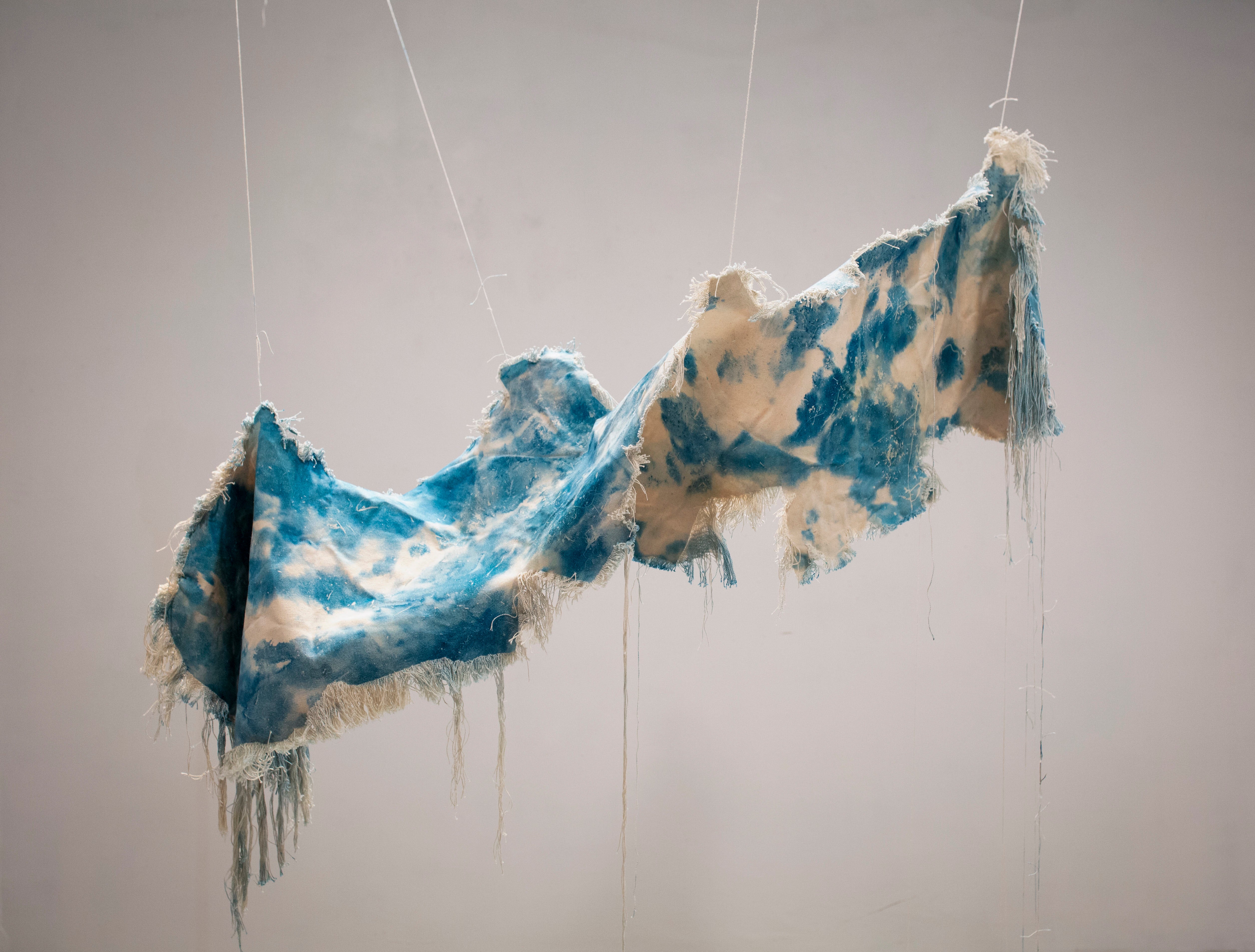 A beige canvas, dyed with spots of cyanotype is twisted and suspended in the air. The edges of the canvas have been unraveled and hand downward. 