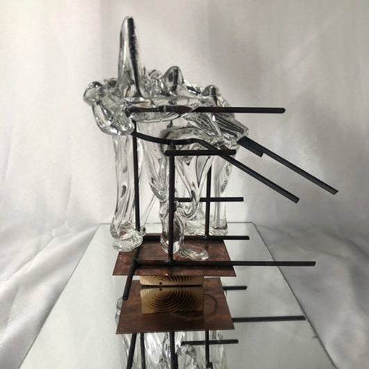 color photograph of a glass and metal sculpture sitting on a mirror. The glass flows over the metal bars of the sculpture.
