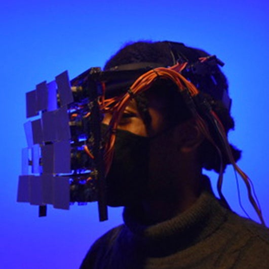 Color photo of a young black person wearing a wearable mechanical sculpture over their face.