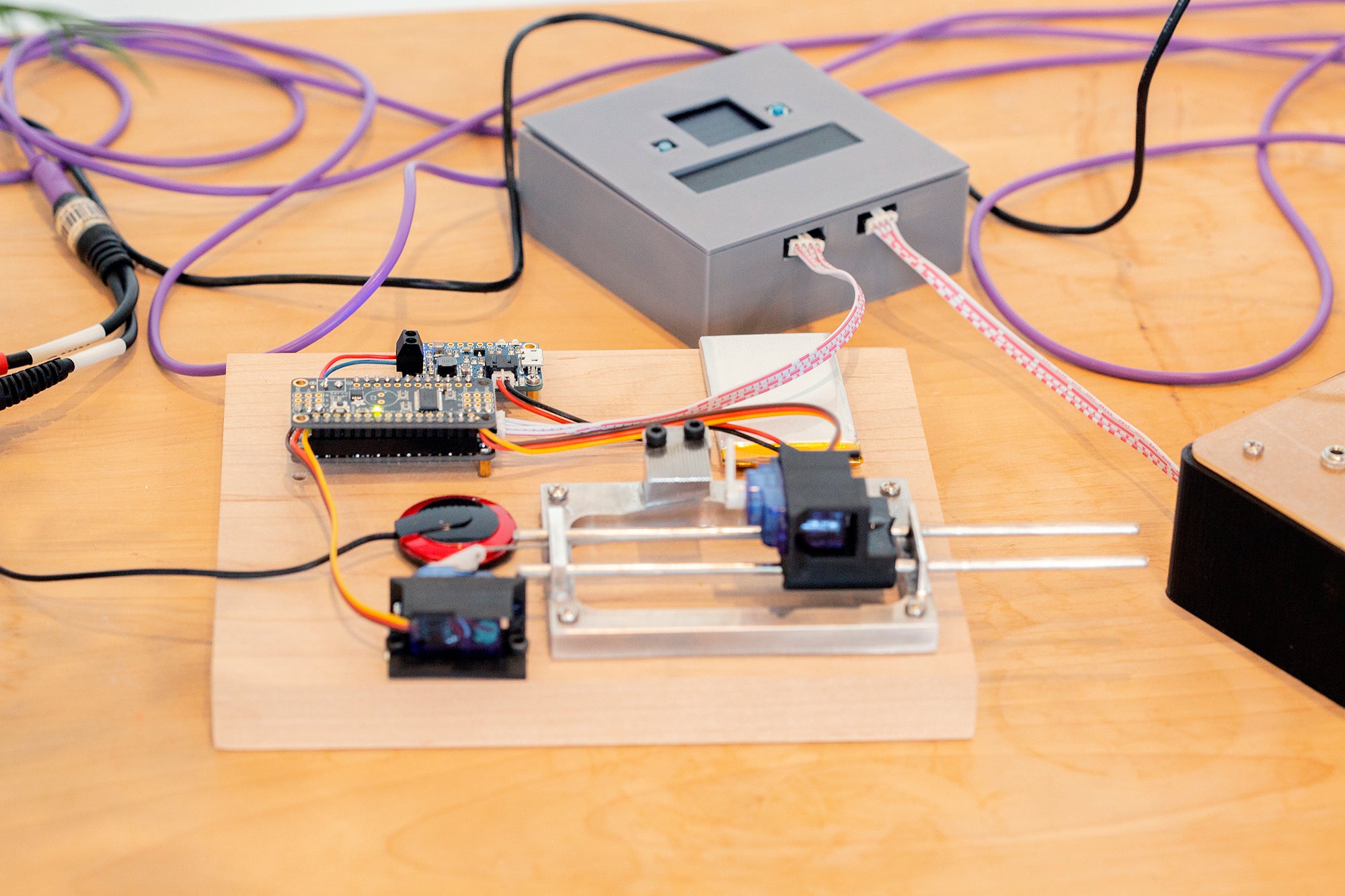 A close view of a robotic music box. A small motorized plastic lever is angled upward facing the music box tines. In the background, a small gray box can be seen. Wires connect the box and a circuitboard on the robotic music box.