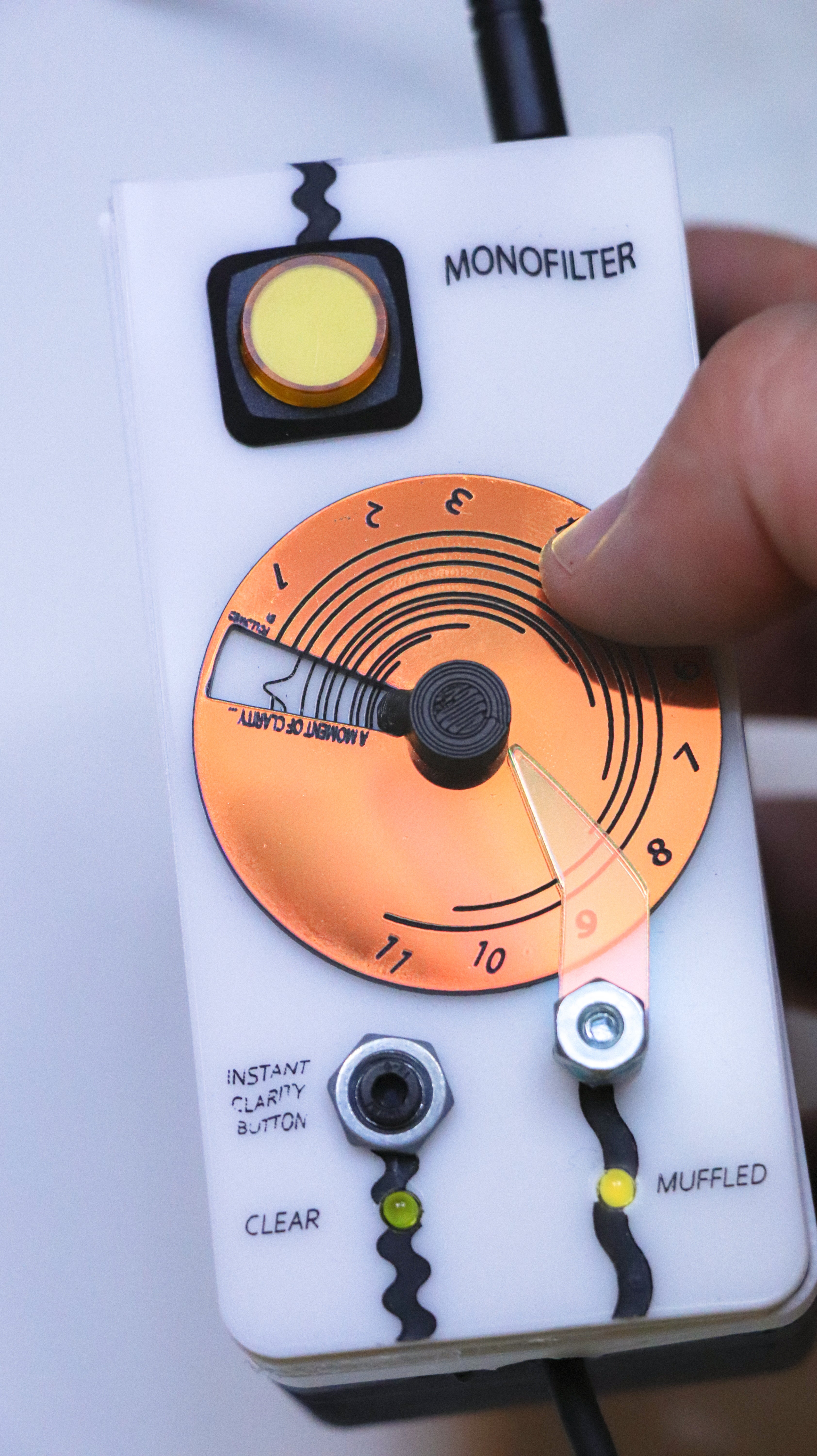 photograph showing a hand holding the Monofilter (roughly the size of a remote, but deeper). The top left corner has a large toggle-button (on/off), the center has a large flat circle that rotates: a dial to change the "muffling duration" (an time interval between each moment of musical clarity and the next). below the dial is an "instant clarity" button, and two lights to indicate whether the sound is "clear" or "muffled". A thumb is placed on the dial.