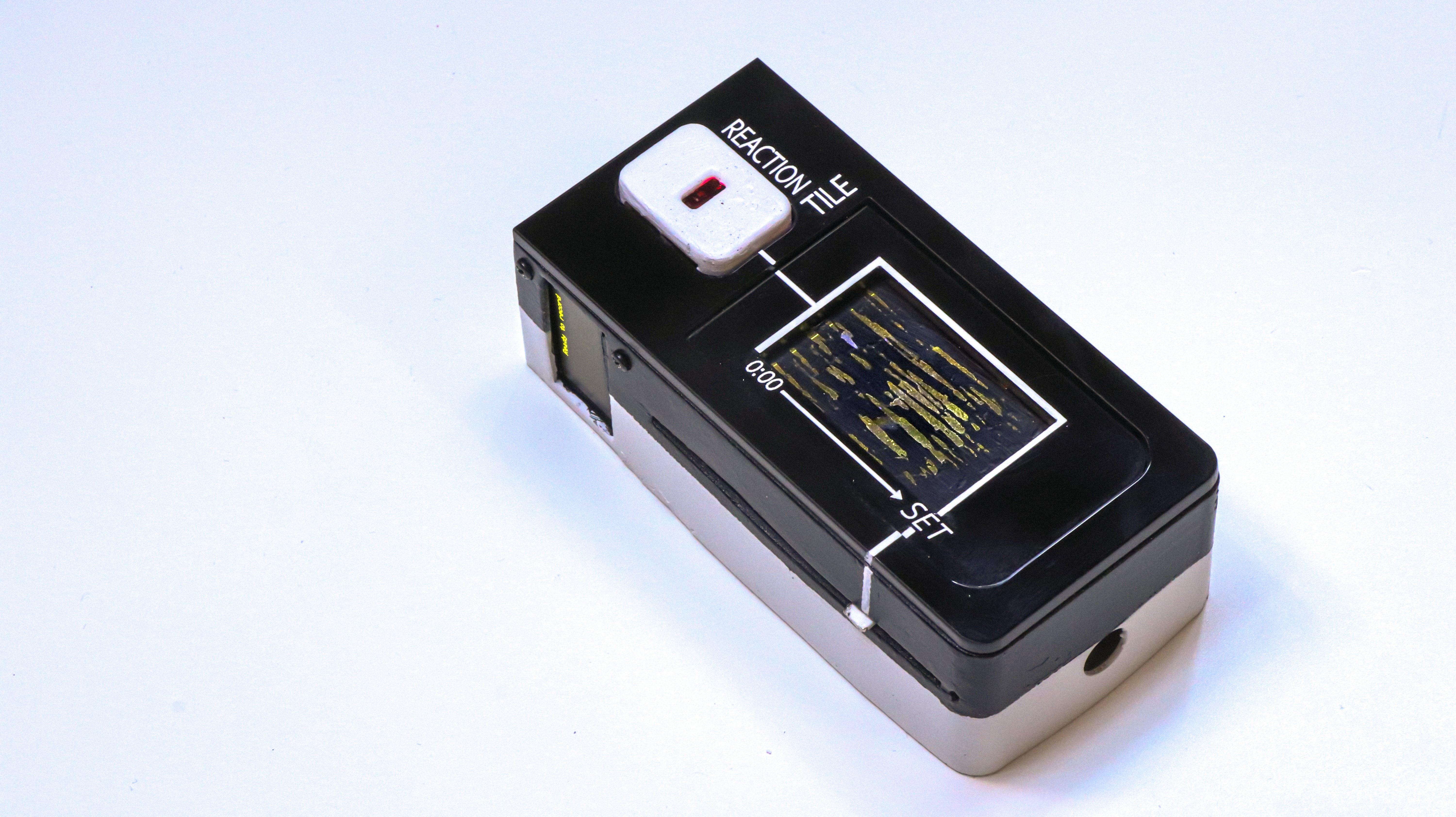 Photograph of the reaction tile: a device about the size of a remote but thicker (1.5inches total). The top surface has a heart rate sensor located near the top (for the thumb). The bottom 2/3 of the top surface is occupied by a flap (like casette players) to place the tile in. The side has a small screen and a slider control to set the total duration to sync the engraving speed/ song duration.