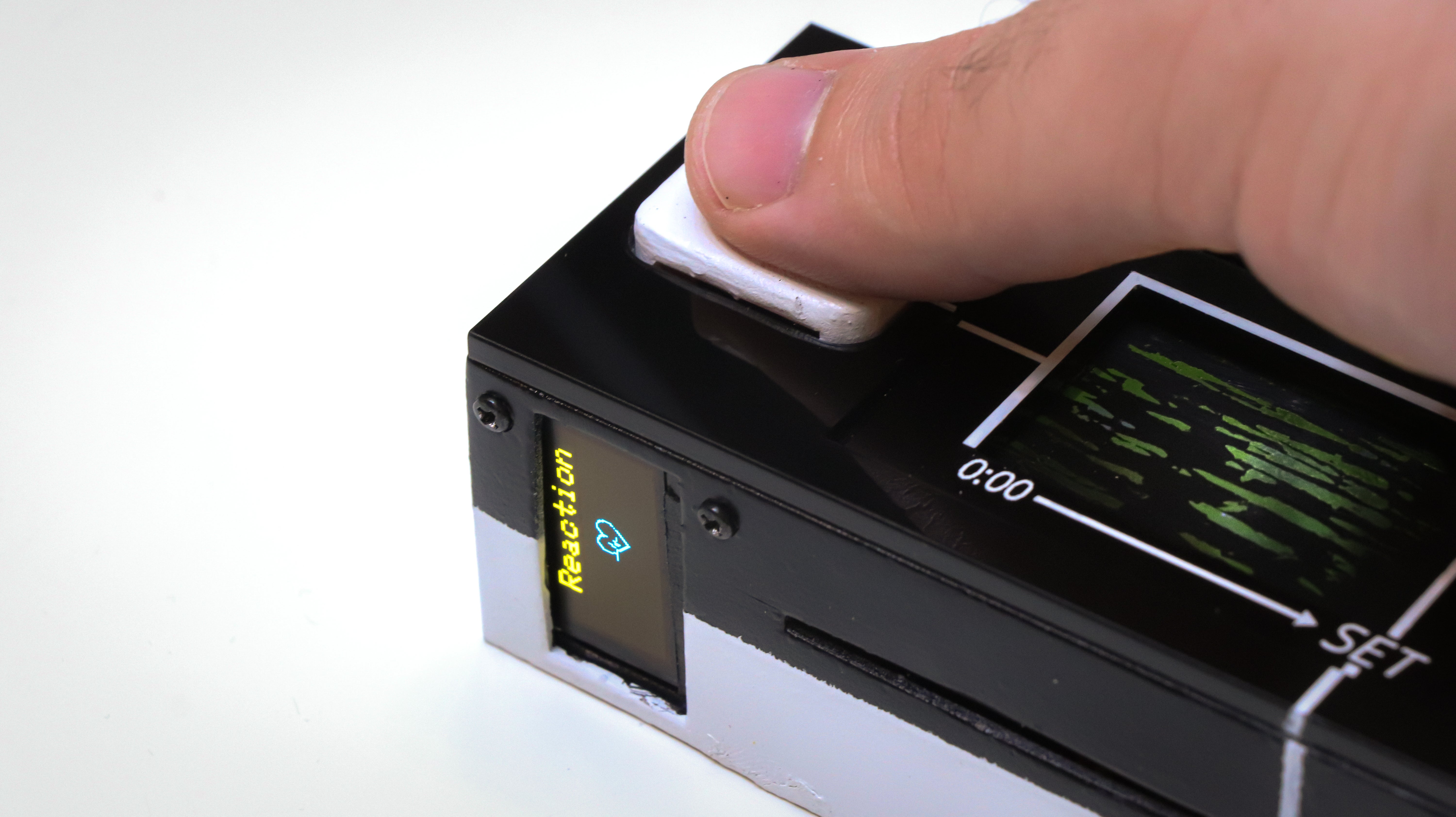 Closeup photograph showing the Reaction Tile (device, roughly the dimensions of a remote but about an inch and half thick) held by a hand. The thumb is on a heart rate sensor (top surface), below is where the tile is placed for engraving: the device opens up like a cassette player. A screen on the side has a small screen showing "reaction" and a cartoon heart.