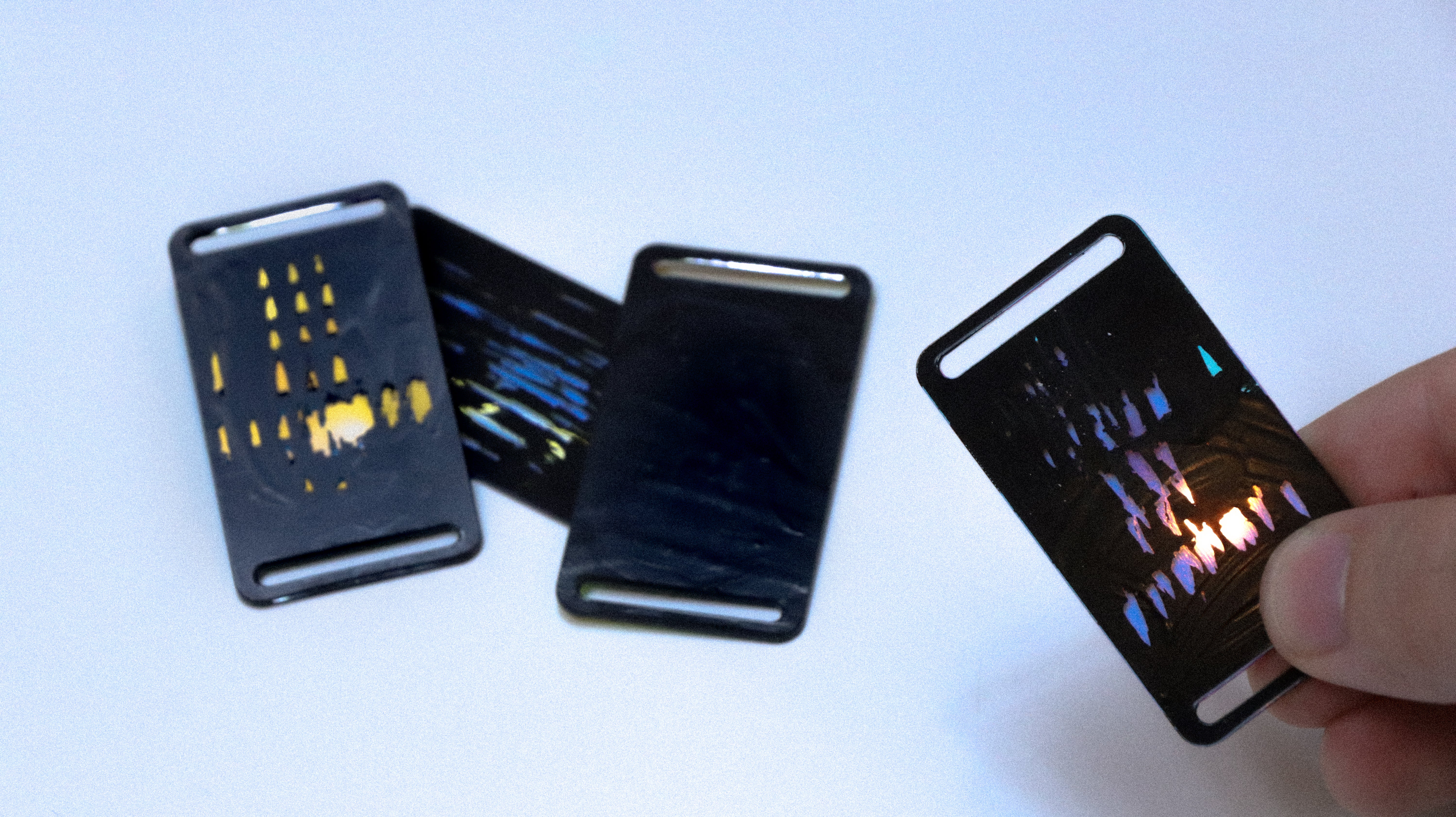 Photograph showing three (domino-sized but 1/8 inches thick) "tiles", each is black (thin layer of paint) but with visible light reflections underneath, as a result of engraving that scratched off some paint in reaction to heart beat variability. On the right side of the image is a hand holding up a tile. bordering the thinner edge of each tile is a thin (0.1in) slot that stretches along; this provides ways to secure tiles to other things (eg a frame, tied to a string, shoelaces?)