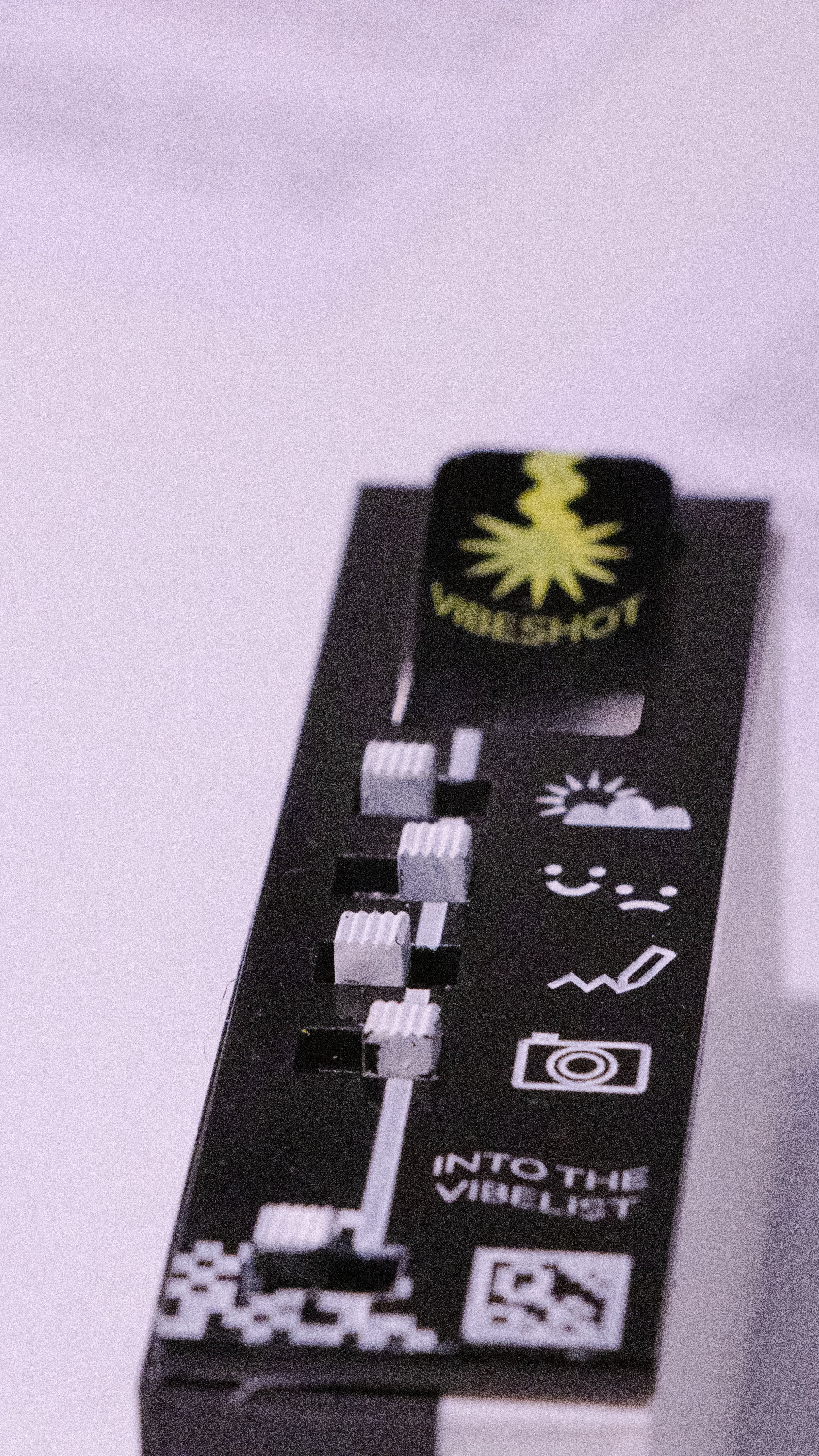 Photograph showing the Vibeshot feature: which is on the side surface of the device. The top of this face has a button labeled "vibeshot". From the button stretches a line which is interrupted by five toggle switches; symbolizing whether the elements the toggles represent are capture in the vibelist. the elements (top to bottom) are weather+location, self-reported feeling, text, image and a web-link (to share with others)