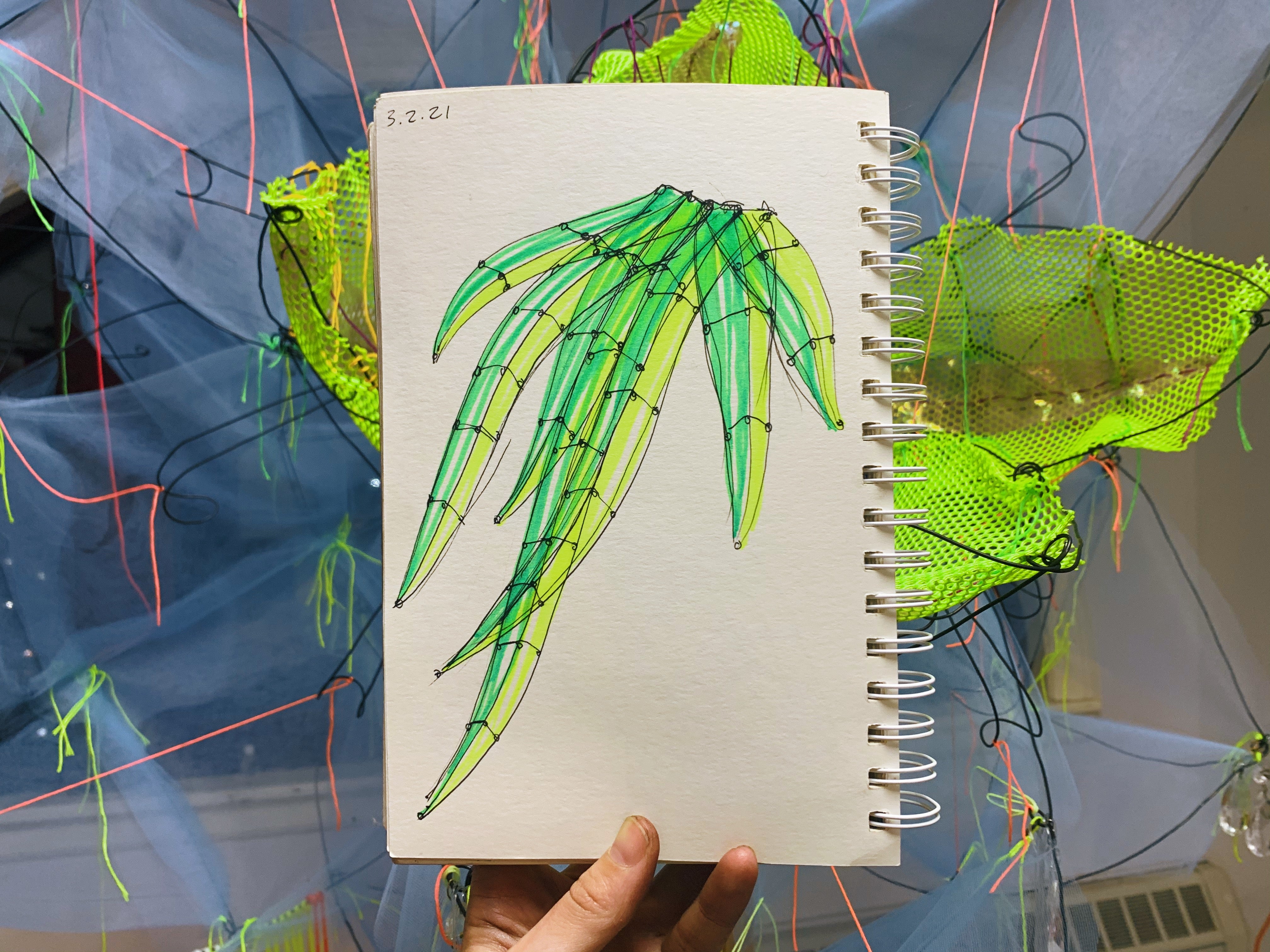 Sketch of how blades of grass can be turned into a wire sculpture. Page held by hand in front of a wire and fabric sculpture of a magnolia. - The Ephemeral Altar & The Floating Grave - Hannah Suzanna