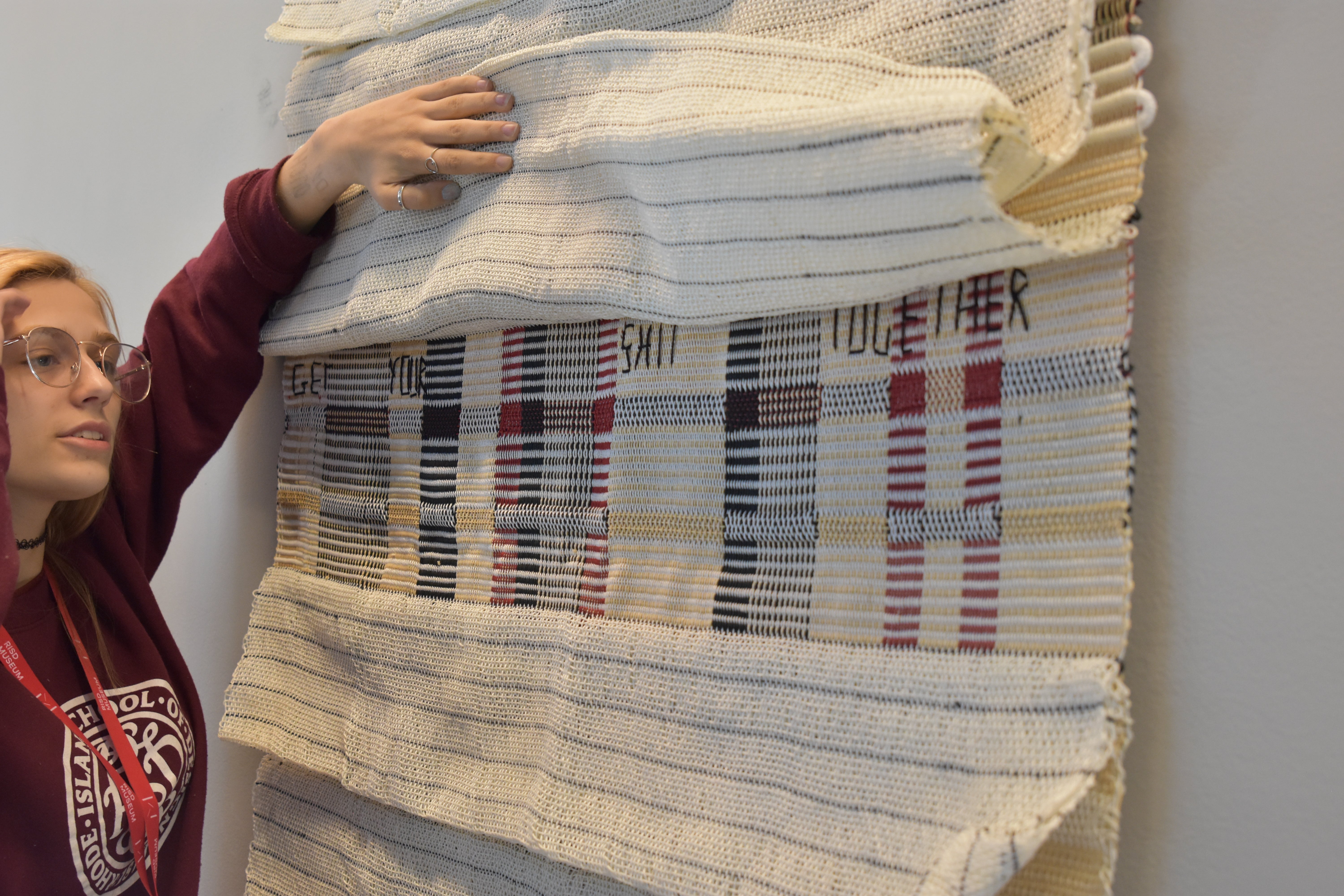 A young woman in a RISD sweatshirt lifts the fold of a hanging woven textile with several flaps