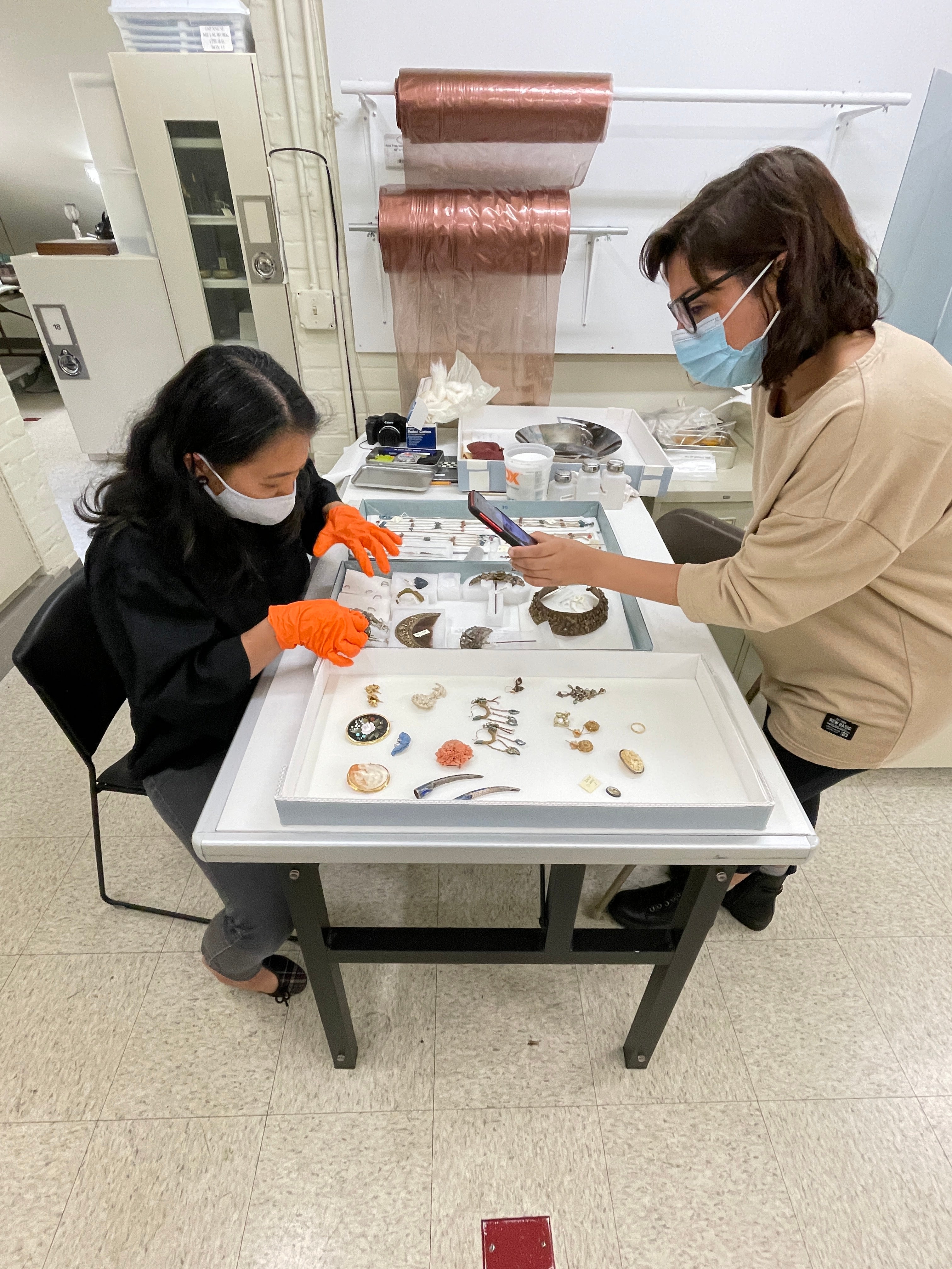 /Two%20masked%20people%20work%20at%20a%20table%20together.%20They%20examine%20trays%20of%20small%20objects%20set%20between%20them.