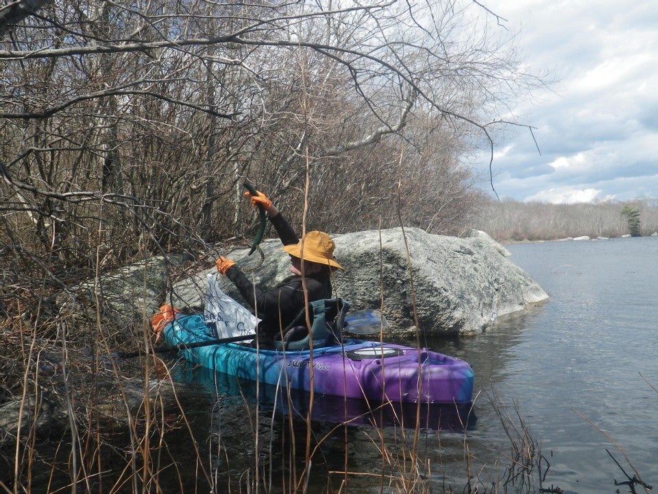 /A%20person%20seated%20in%20a%20kayak%20on%20calm%20water.%20The%20trees%20above%20them%20and%20the%20vegetation%20we%20view%20them%20through%20are%20leafless.%20The%20sky%20is%20cloudy.