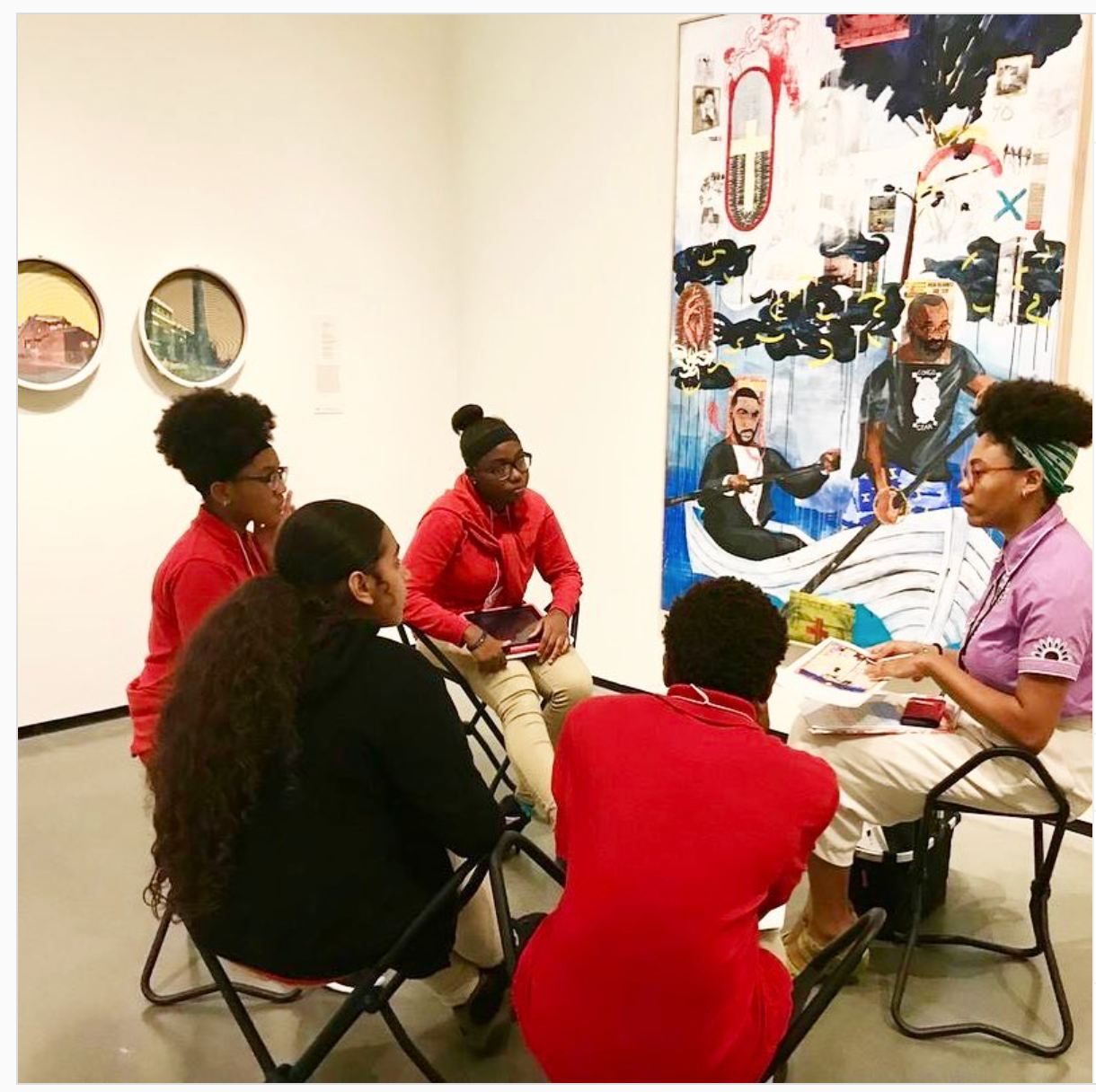 /Four%20young%20dark-skinned%20people%20engaged%20in%20conversation%2C%20sitting%20in%20front%20of%20a%20dynamic%20abstract%20painting%20of%20two%20dark-skinned%20people%20rowing%20a%20boat.