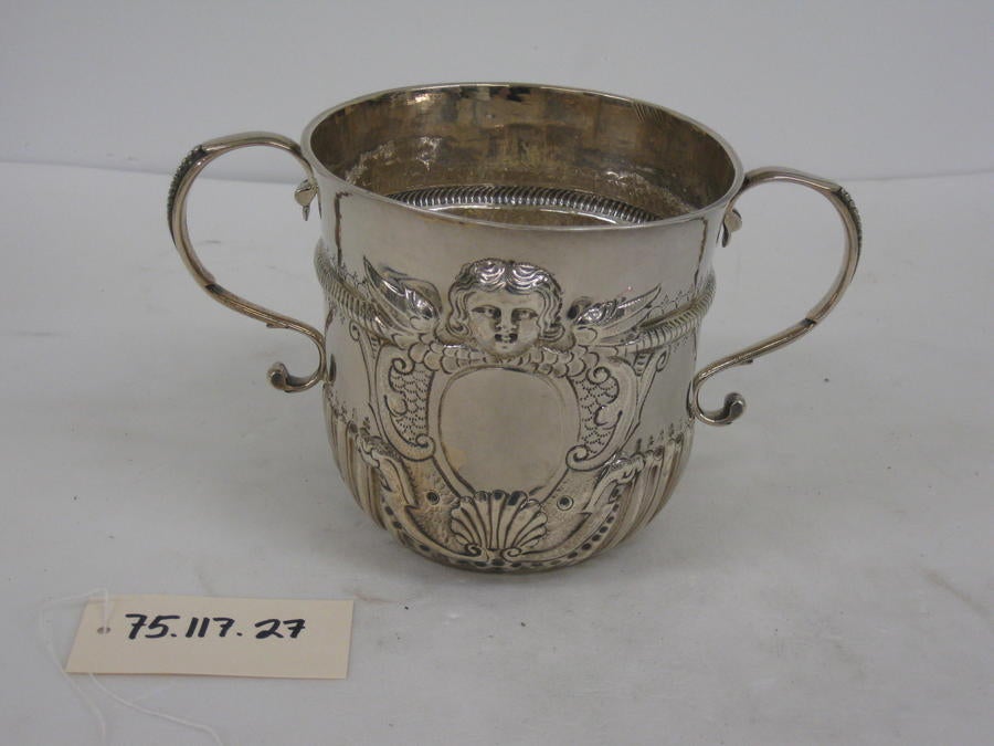 /A%20silver%20cup%20with%20symmetrical%20handles%2C%20swirling%20raised%20designs%20and%20a%20small%20raised%20face%20on%20the%20upper%20half%20of%20the%20cup.