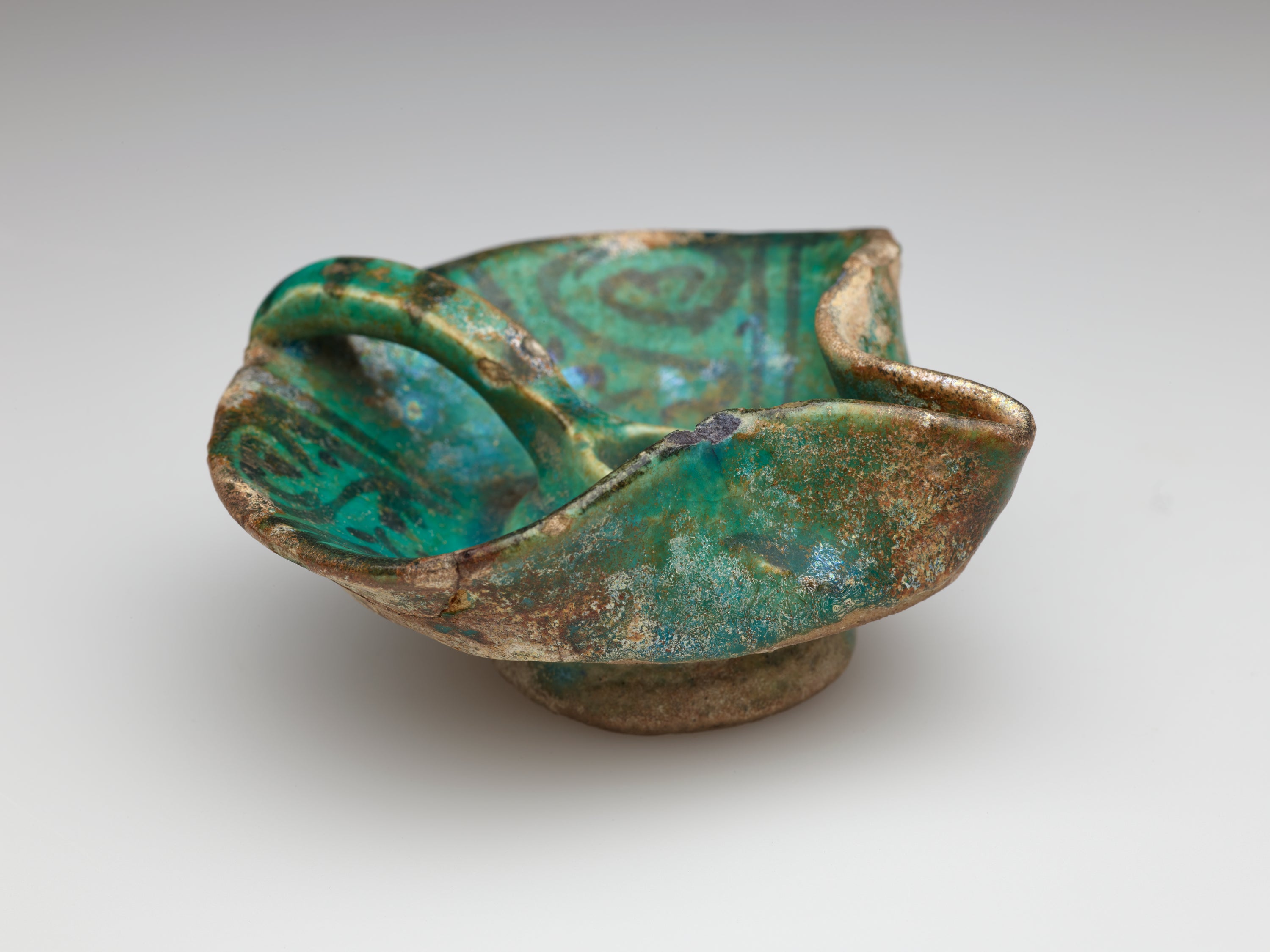 /An%20irregularly%20shaped%20blue-green%20iridized%20vessel%20with%20dark%20designs.%20Rounded%20on%20one%20side%20with%20a%20handle%20that%20comes%20to%20the%20center%20of%20the%20vessel.%20The%20other%20side%20has%20a%20folded%20spout-like%20edge.