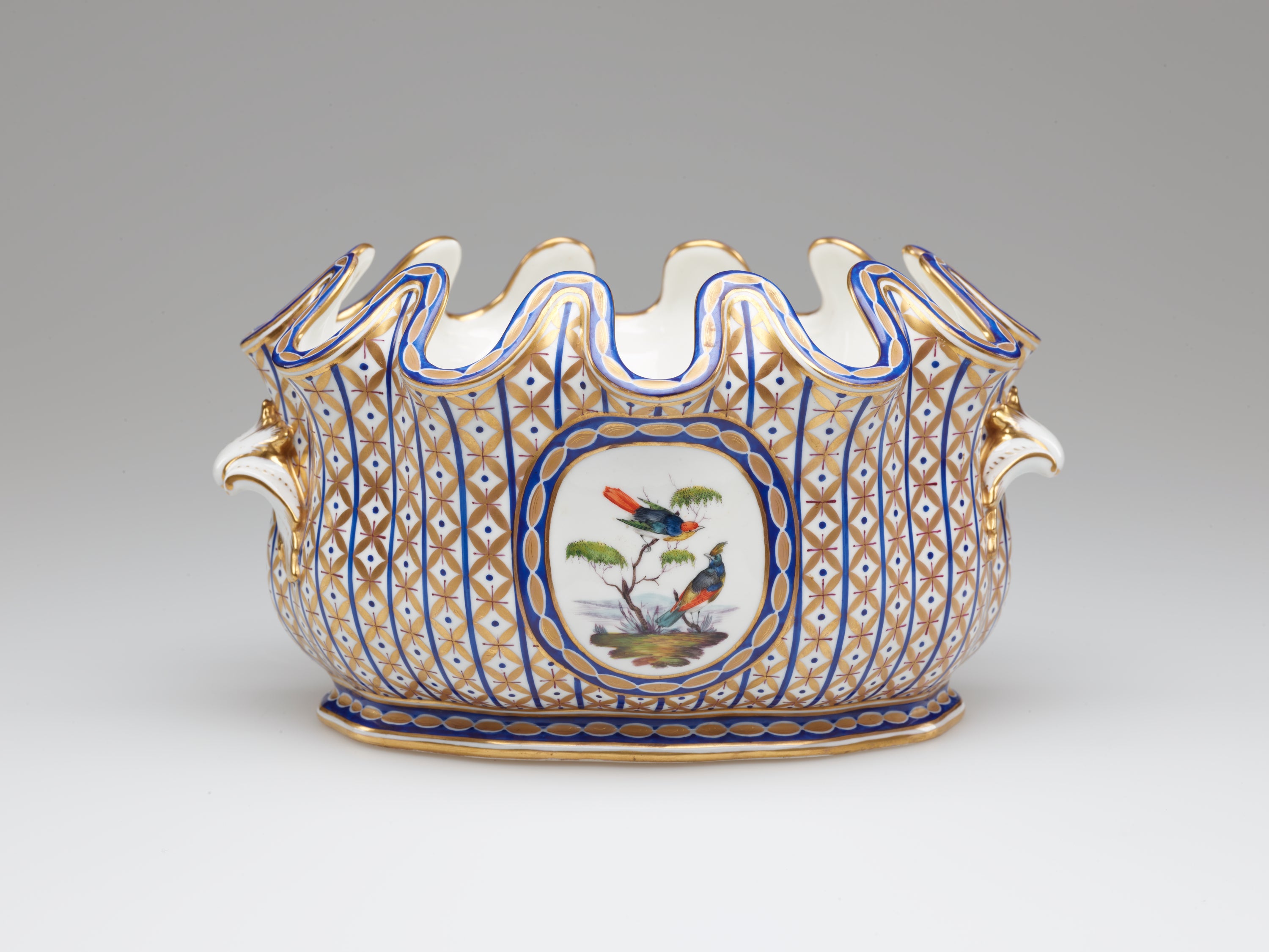/An%20open%20vessel%20with%20a%20deeply%20scalloped%20rim.%20A%20central%20decoration%20of%20two%20birds%20and%20a%20tree%20is%20surrounded%20by%20vertical%20bands%20of%20blue%20with%20gold%20diamond%20shapes.%20
