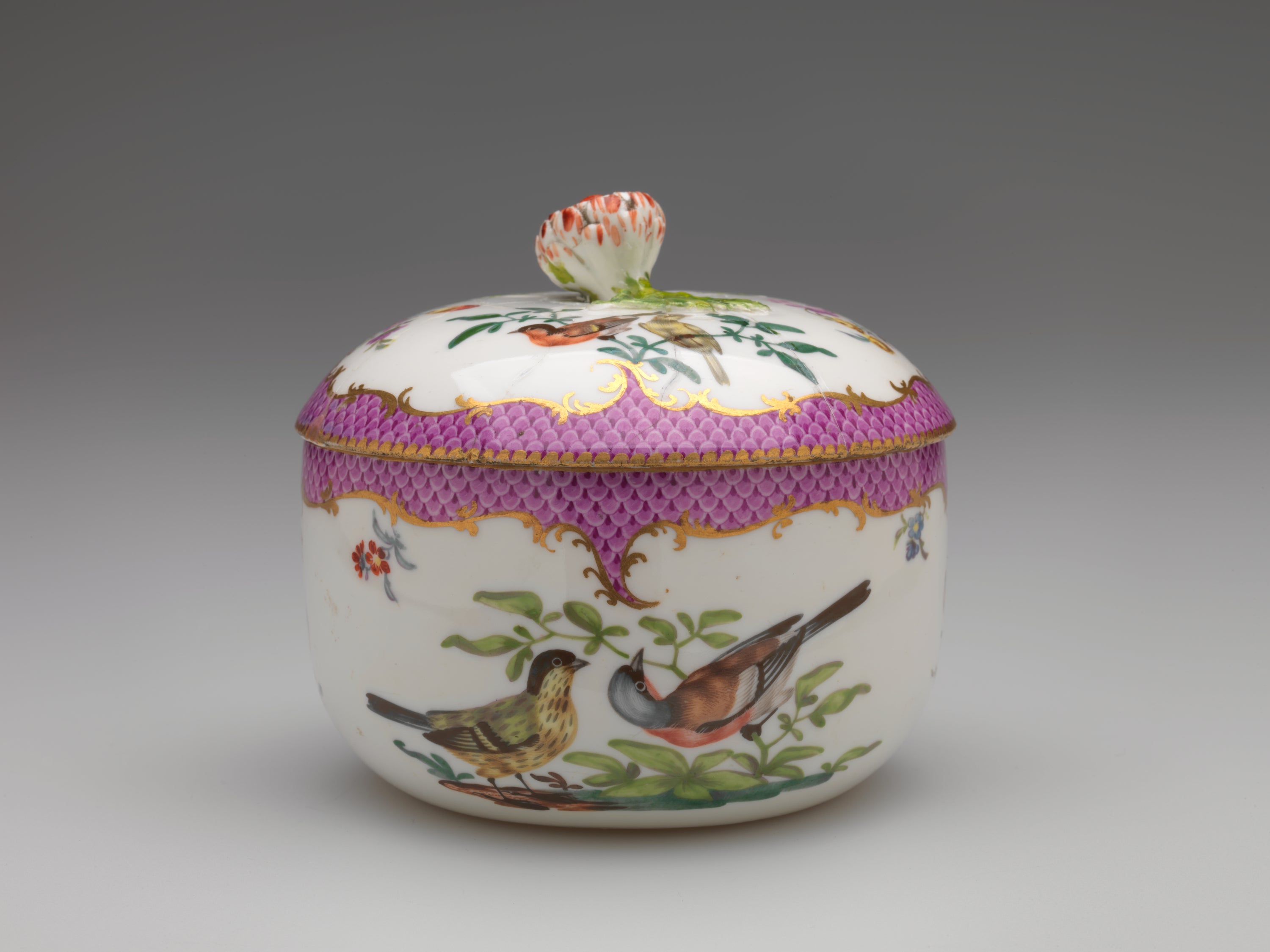 /A%20white%20vessel%20with%20a%20lid%2C%20with%20%20gilded%20and%20pink%20decoration%2C%20floral%20decorations%2C%20and%20two%20birds%20that%20are%20white%2C%20black%2C%20red%20and%20blue%2C%20both%20are%20on%20a%20branch.