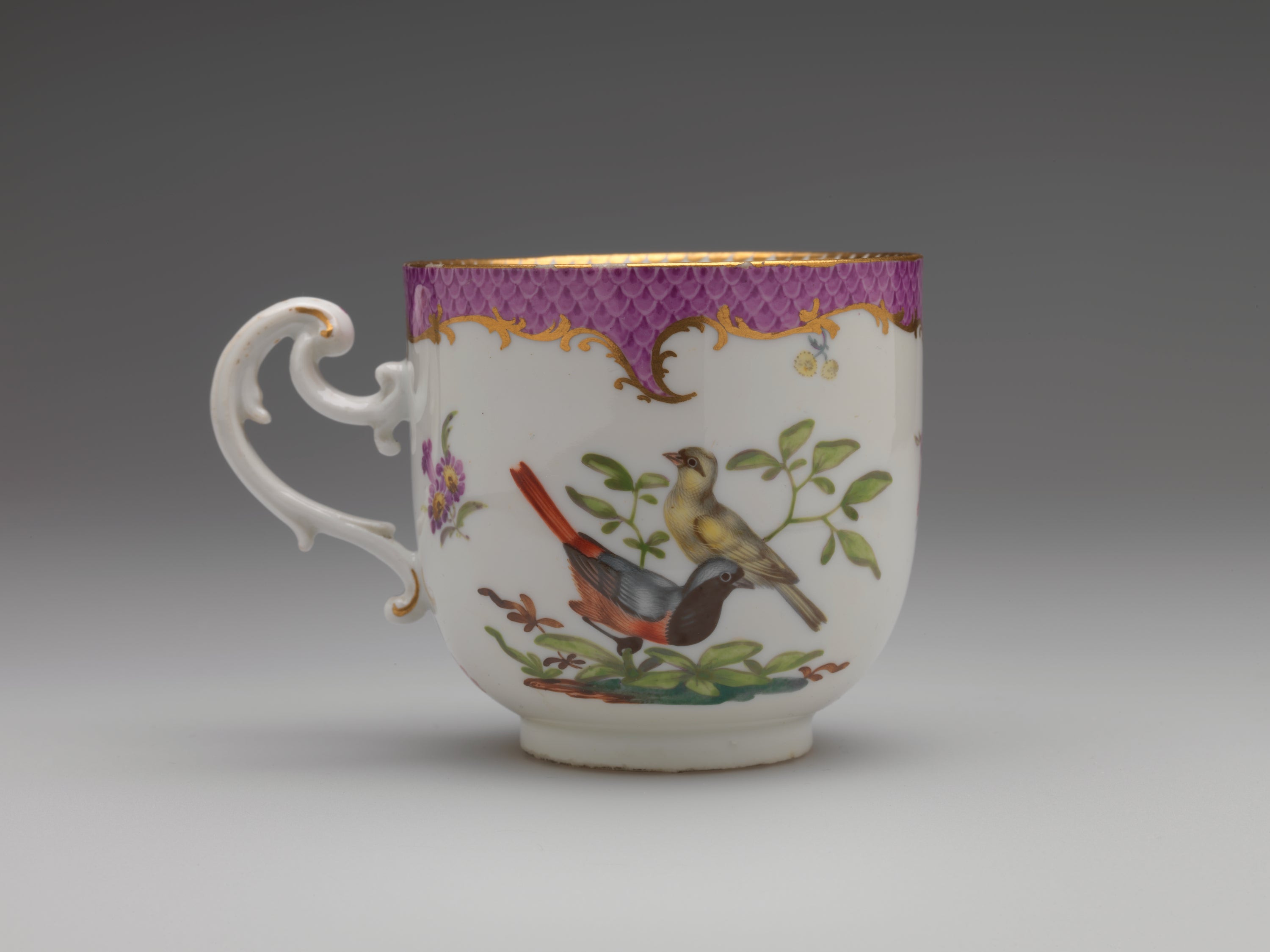 /A%20white%20teacup%20with%20a%20handle%20that%20has%20swirls%2C%20gilding%2C%20pink%2C%20and%20floral%20decorations.%20There%20are%20two%20birds%2C%20one%20large%20and%20yellow%2C%20the%20other%20is%20white%2C%20black%2C%20blue%20and%20orange.