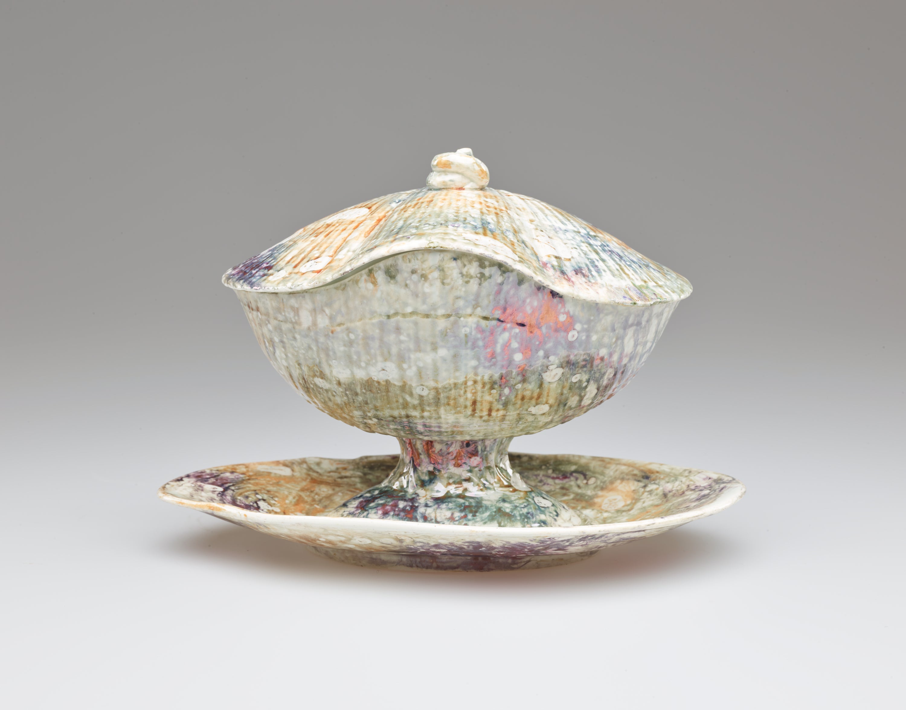 /A%20lidded%20ceramic%20vessel%20on%20an%20organically%20shaped%20tray.%20A%20small%2C%20swirled%20finial%20is%20on%20the%20lid.%20The%20body%20of%20the%20vessel%20has%20ridges%20covered%20with%20a%20mottled%2C%20multi-colored%20glaze.