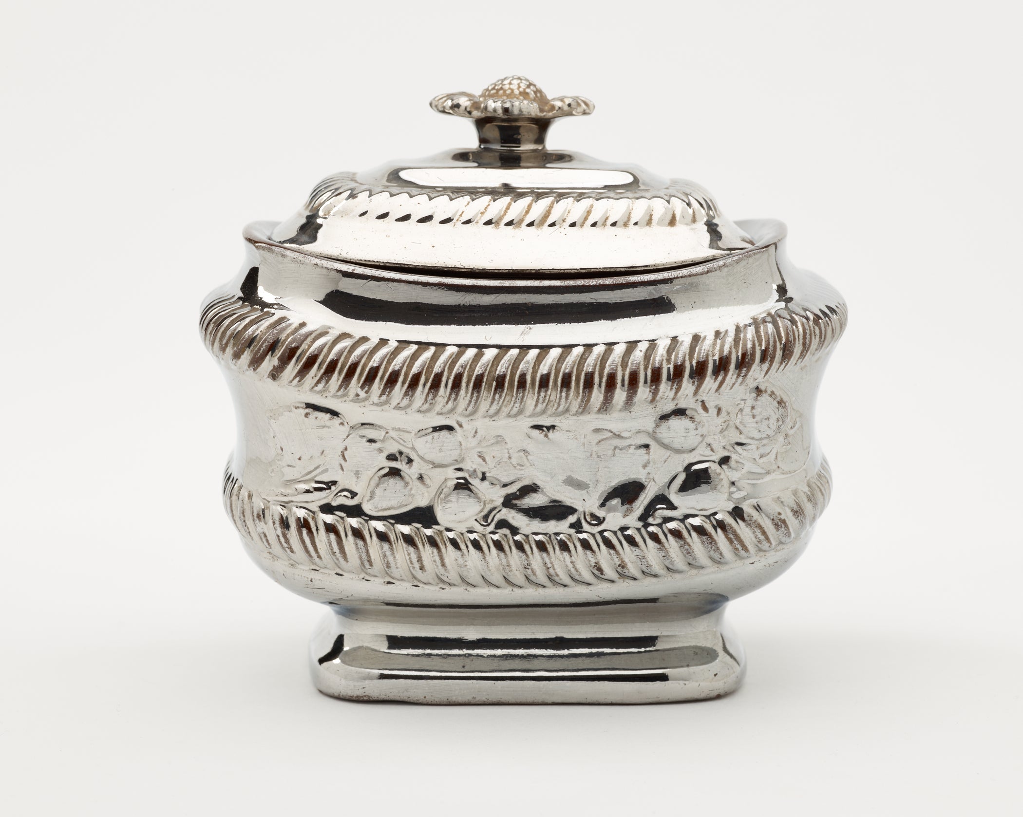 /A%20silver%20luster%20sugar%20bowl%20with%20a%20decorative%20lid%20and%20a%20rounded%20square%20body%20with%20sculptural%20decorations.