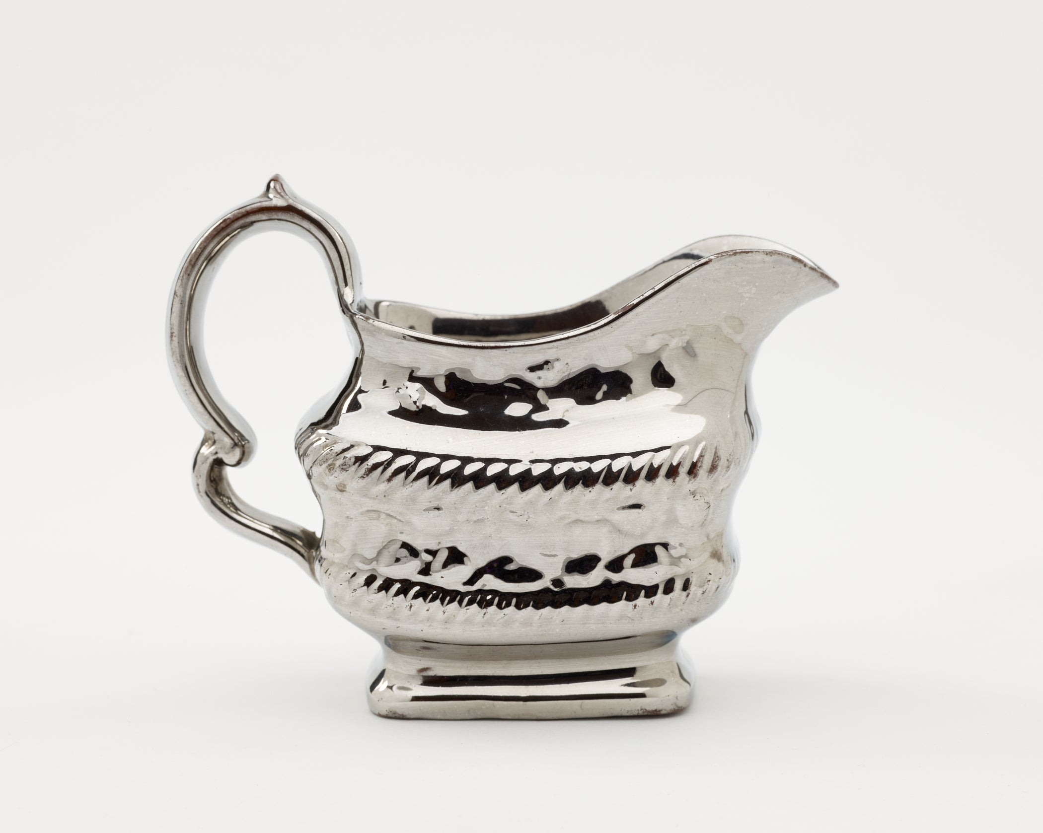 /A%20silver%20luster%20creamer%20with%20a%20decorative%20handle%2C%20a%20rounded%20square%20body%20with%20sculptural%20decorations.