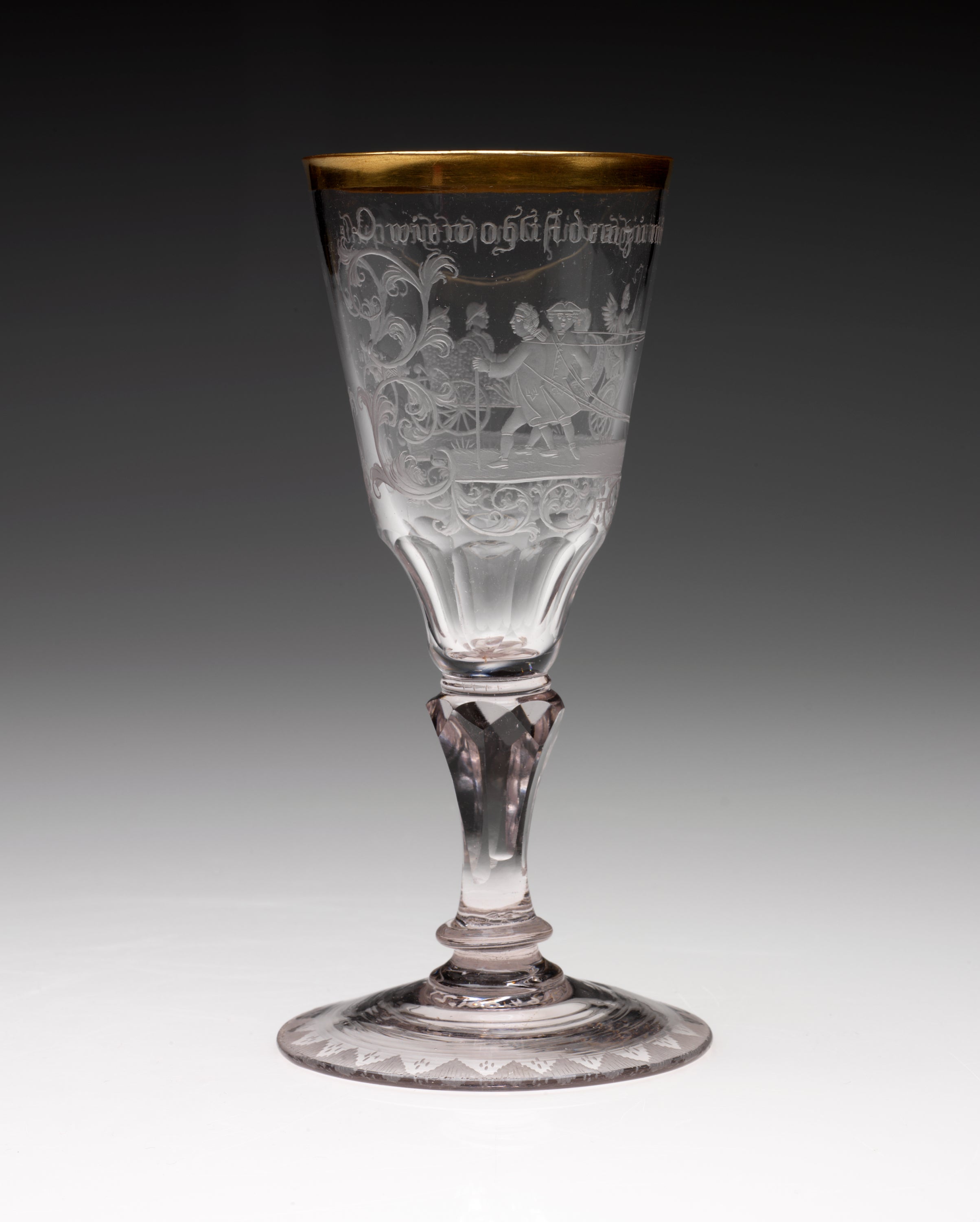 /A%20clear%20stemmed%20glass%20with%20a%20rounded%20foot%20and%20thick%20gold%20rim.%20An%20image%20of%20figures%20walking%20in%20a%20group%20with%20words%20above%20them%20are%20etched%20in%20the%20sides.%20