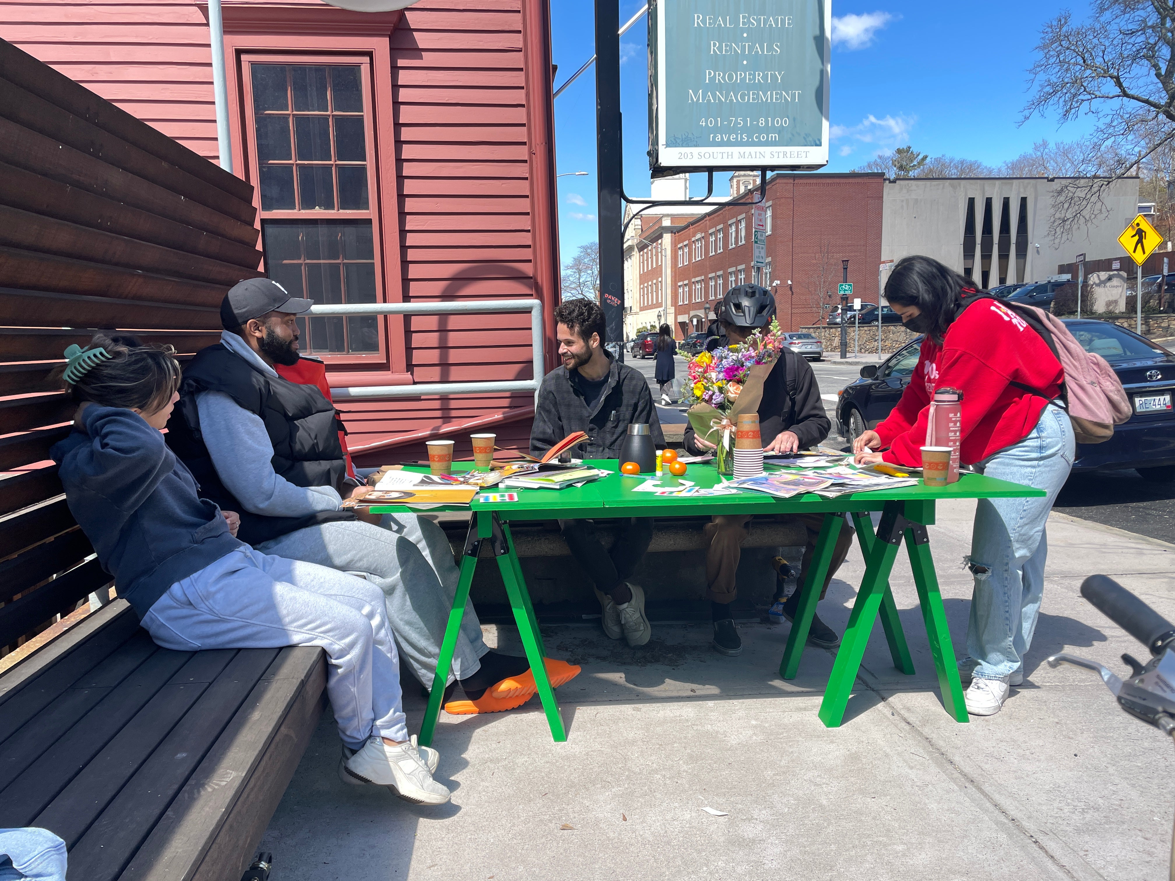 Five students gather around a bright green table that is covered in collage materials, flowers, drinks, and citrus. 
