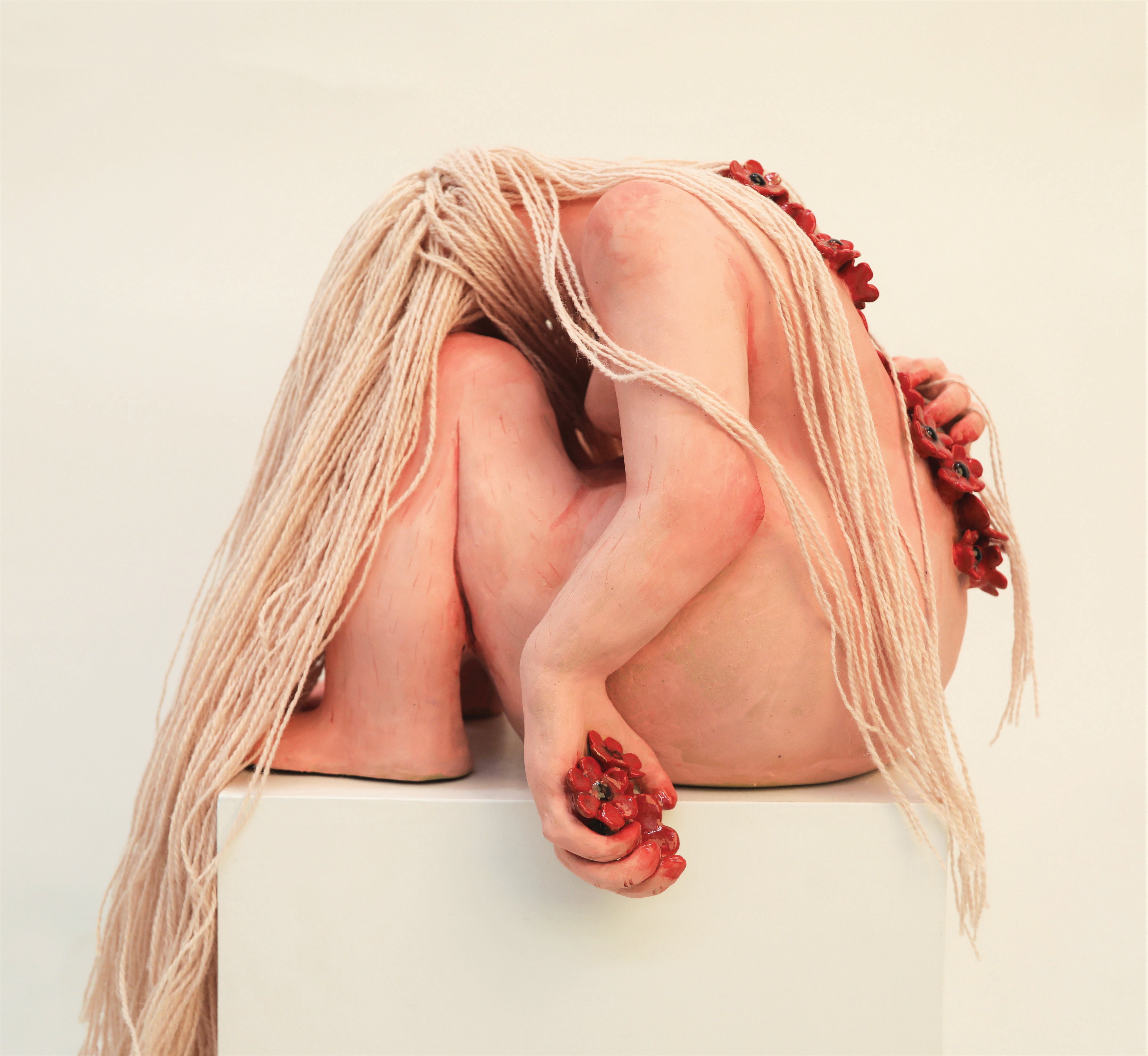 A pink ceramic figure with pink yarn hair rests its head on its knees, looking away.