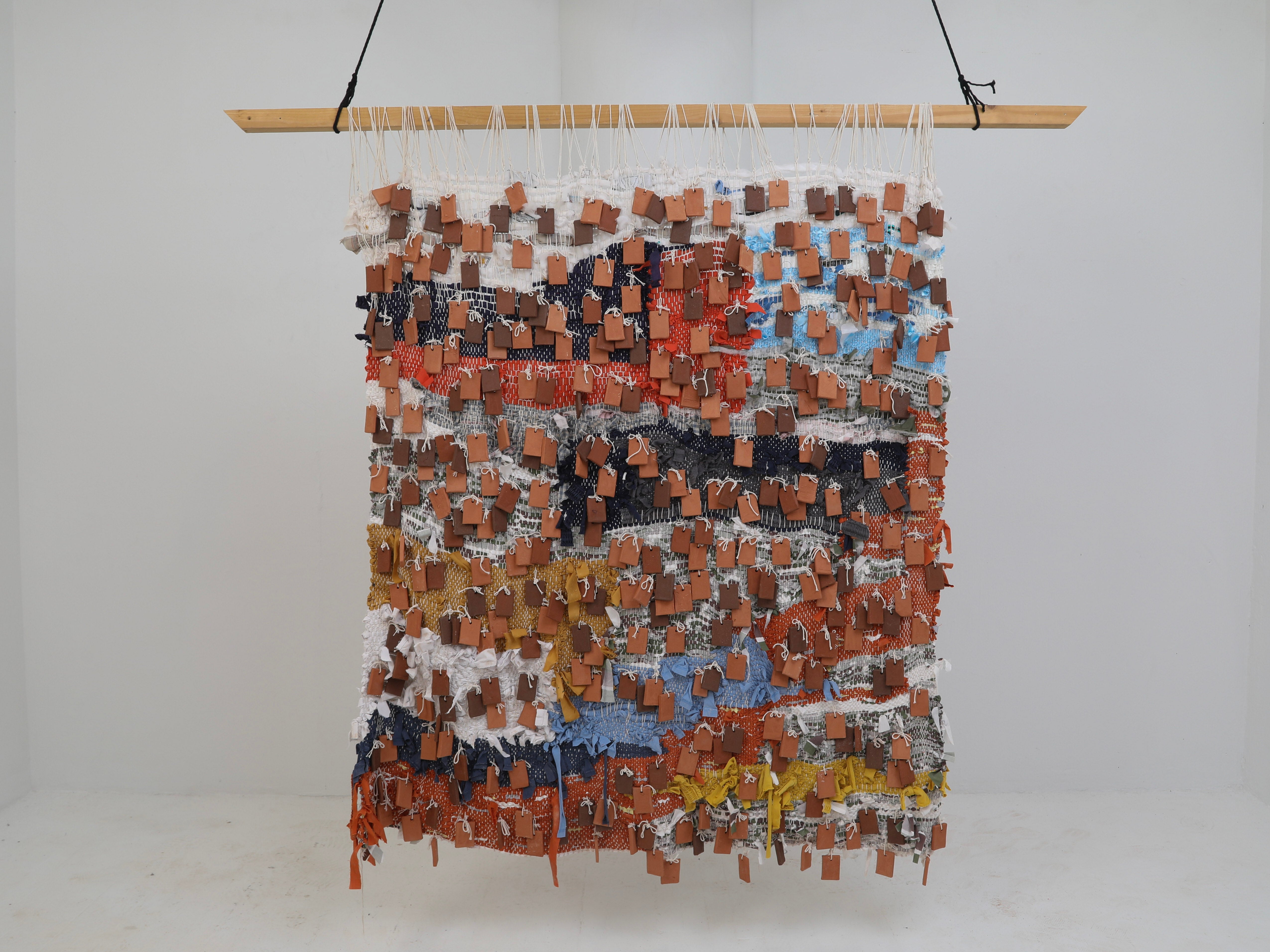 A large, colorful woven textile made of discarded fabrics and old, childhood clothing covered in terracotta tiles hangs