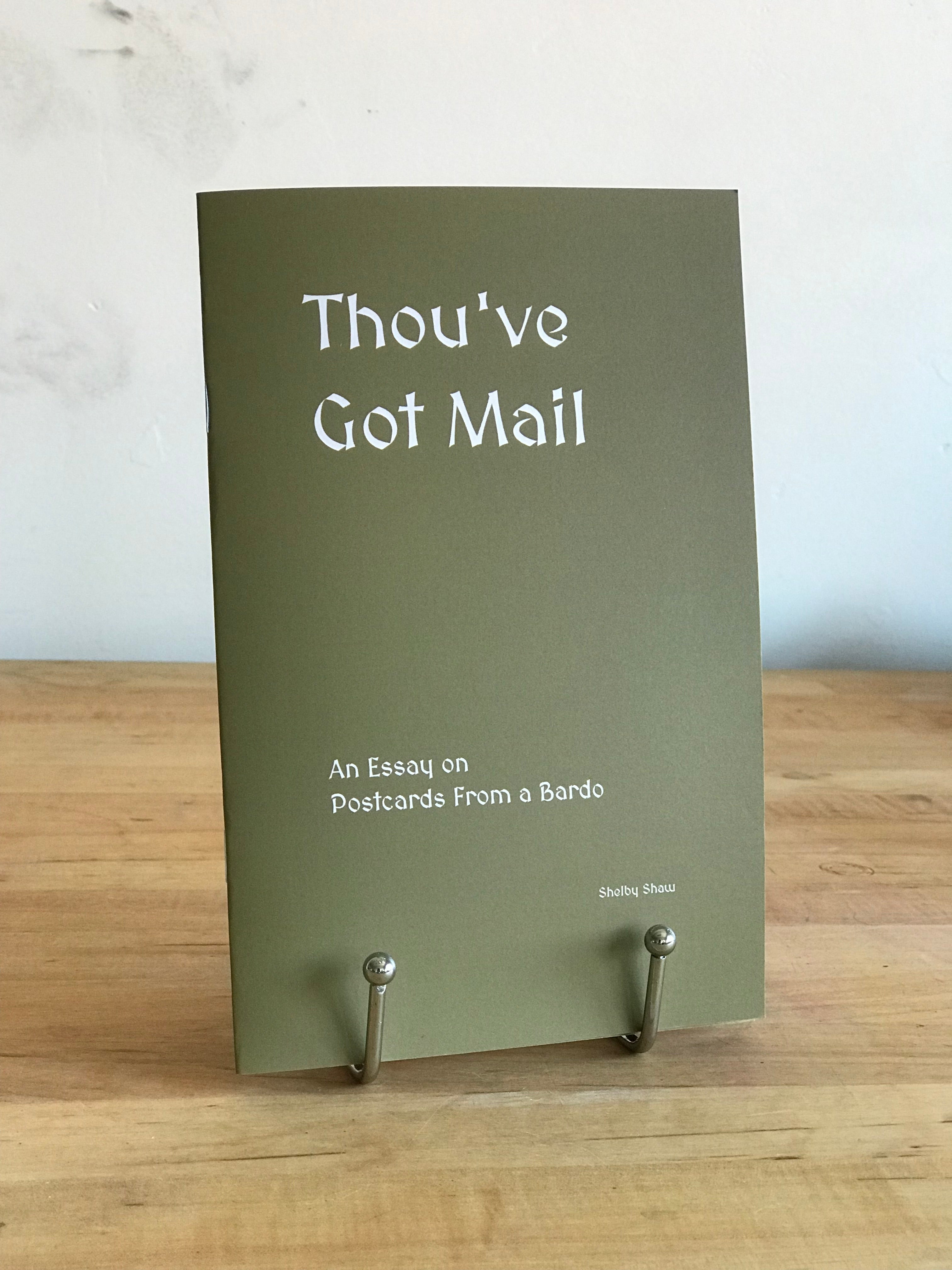 The book 'Thou've Got Mail' which is a khaki green-brown color with white font is displayed on a silver book stand atop a wooden tabletop with a white wall in the background.