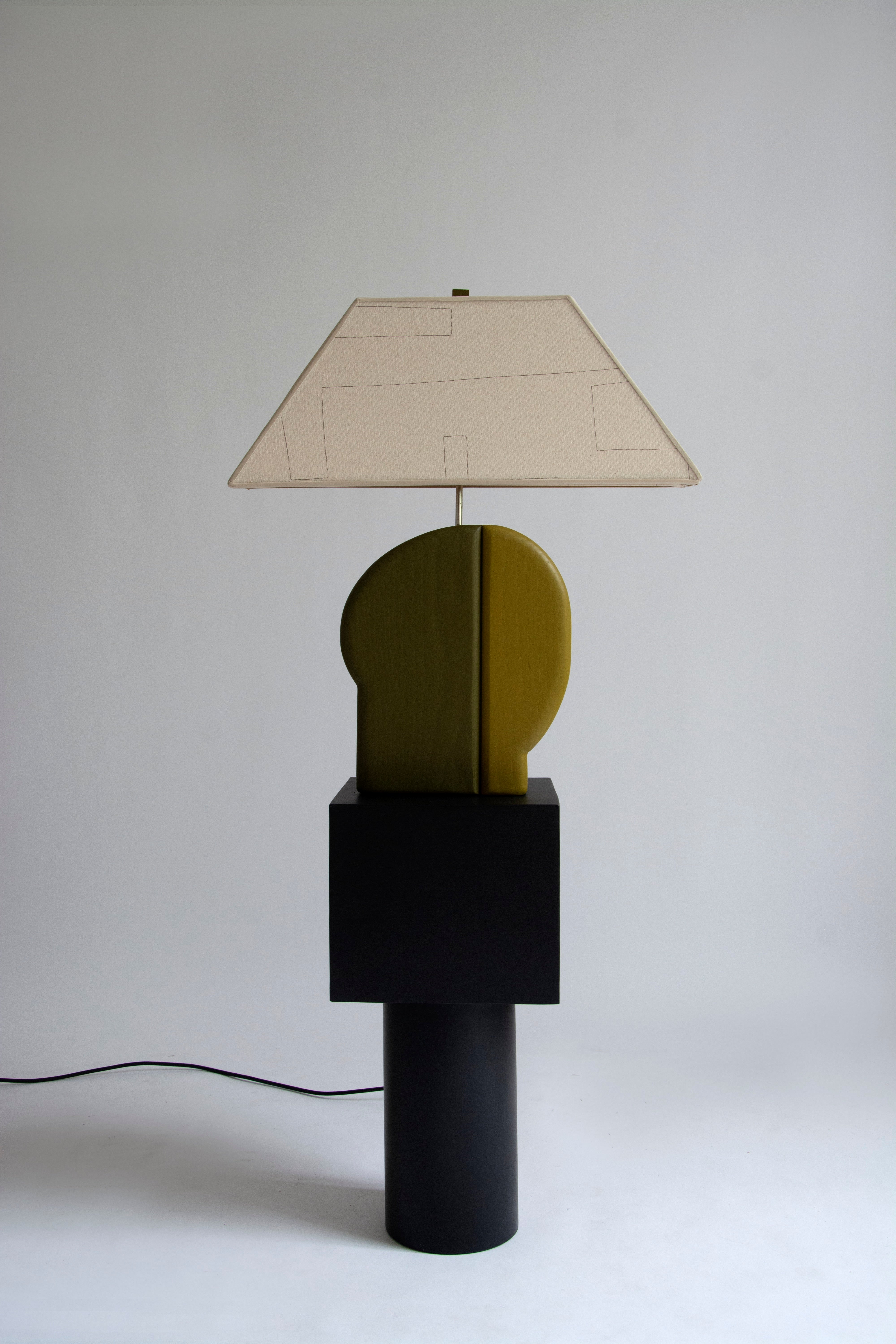 The Side table has a black wooden cylindrical base, with a square black wooden top. A two toned green wooden lamp base and canvas shade with hand sewn lines sits atop the table.  