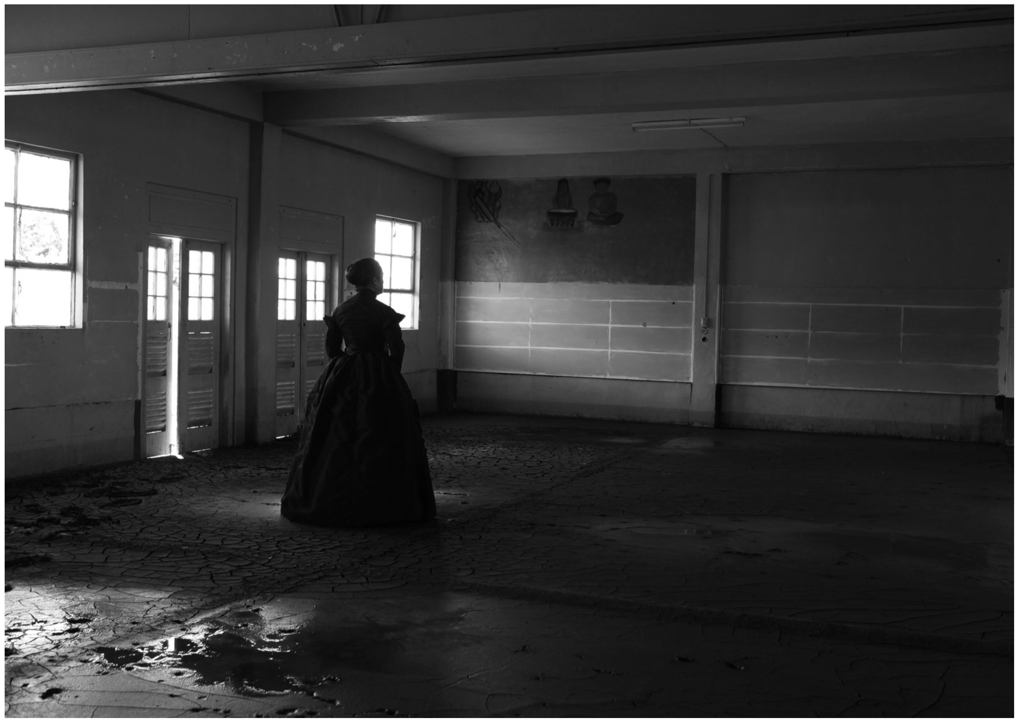 Yuki Kihara's photograph Marist Brothers Old Pupil’s Association House After Cyclone Evan, Mulivai is a black and white indoor scene where a female presenting person in a black Victorian style gown faces an empty wall of an abandoned and poorly lit room.