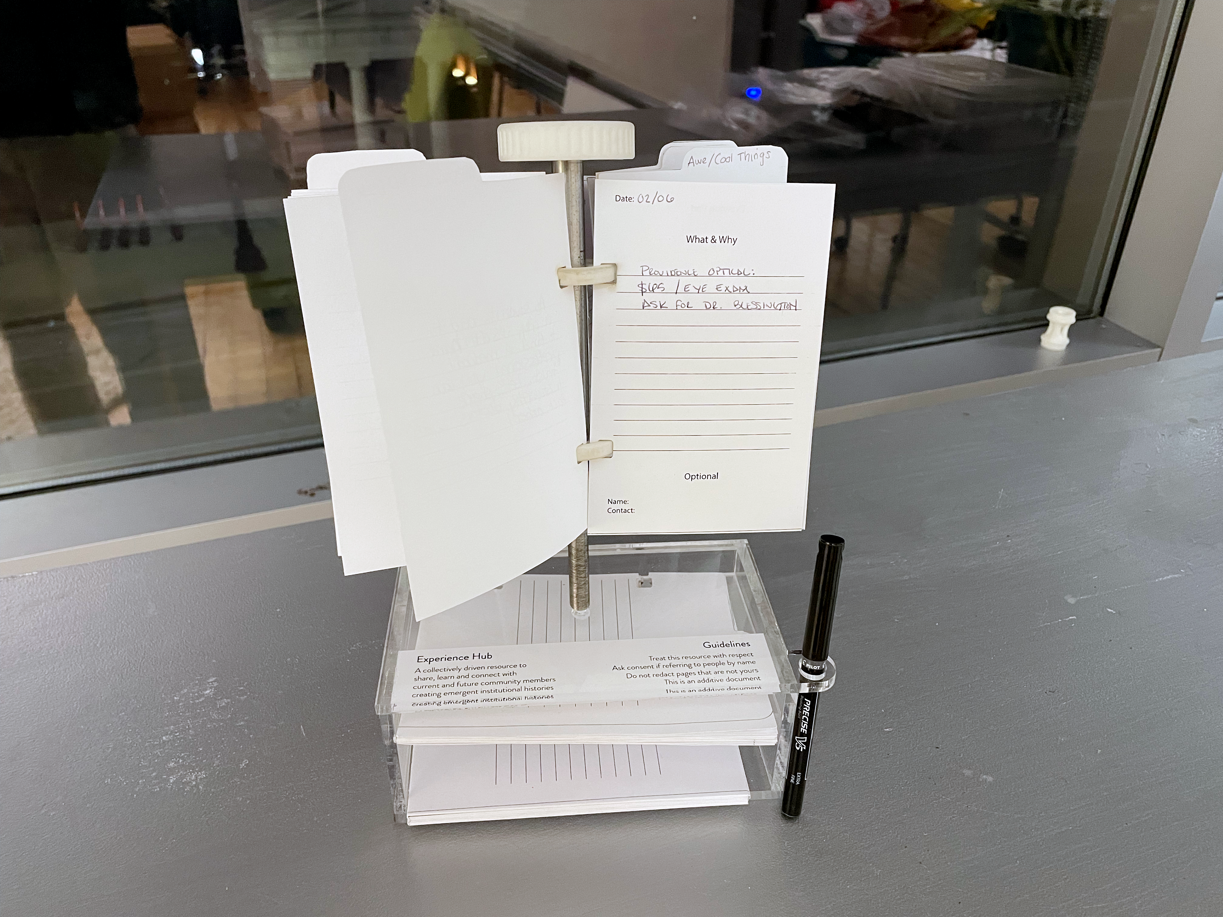 A small steel pole holding half sheets of paper that people have written on. The pole is on top of an clear box holding extra paper and a pen. 