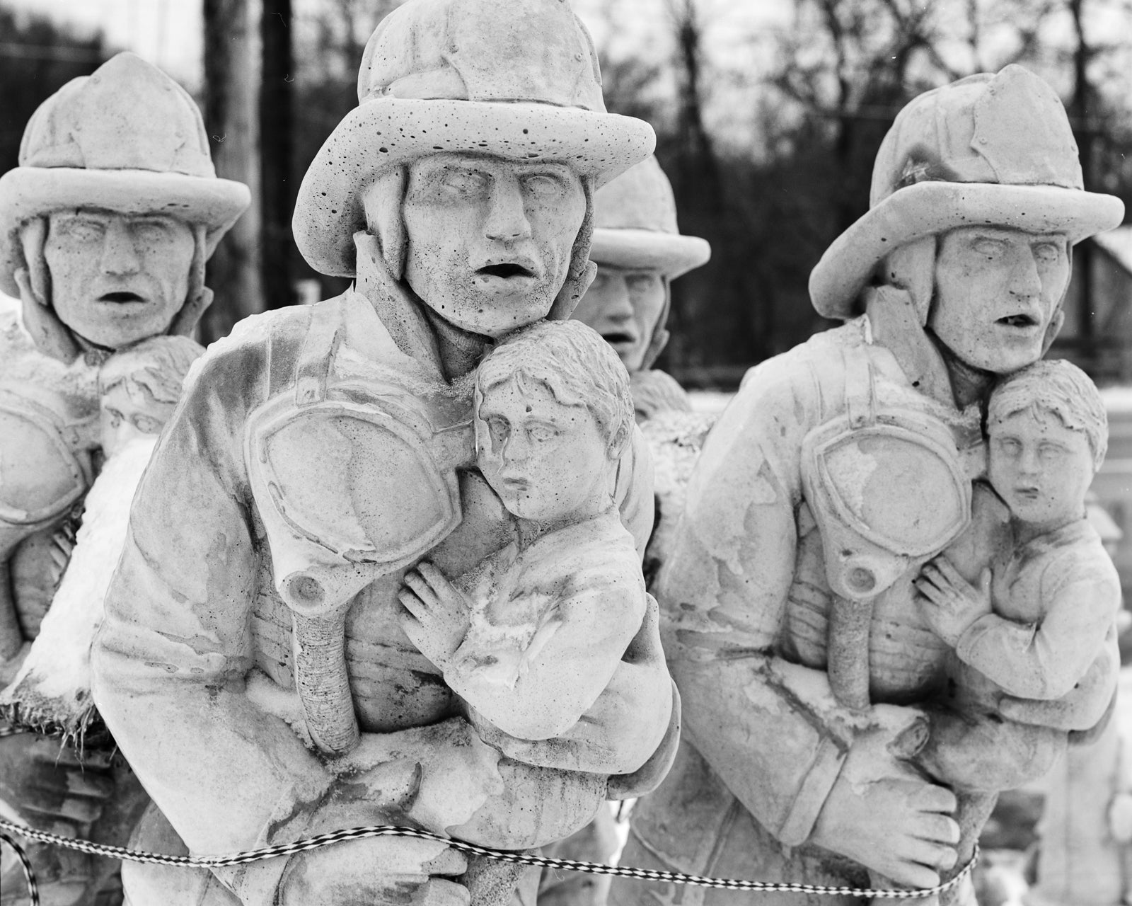 A group of identical white statues of firefighters carrying a child. A rope at the bottom of the frame binds them together.