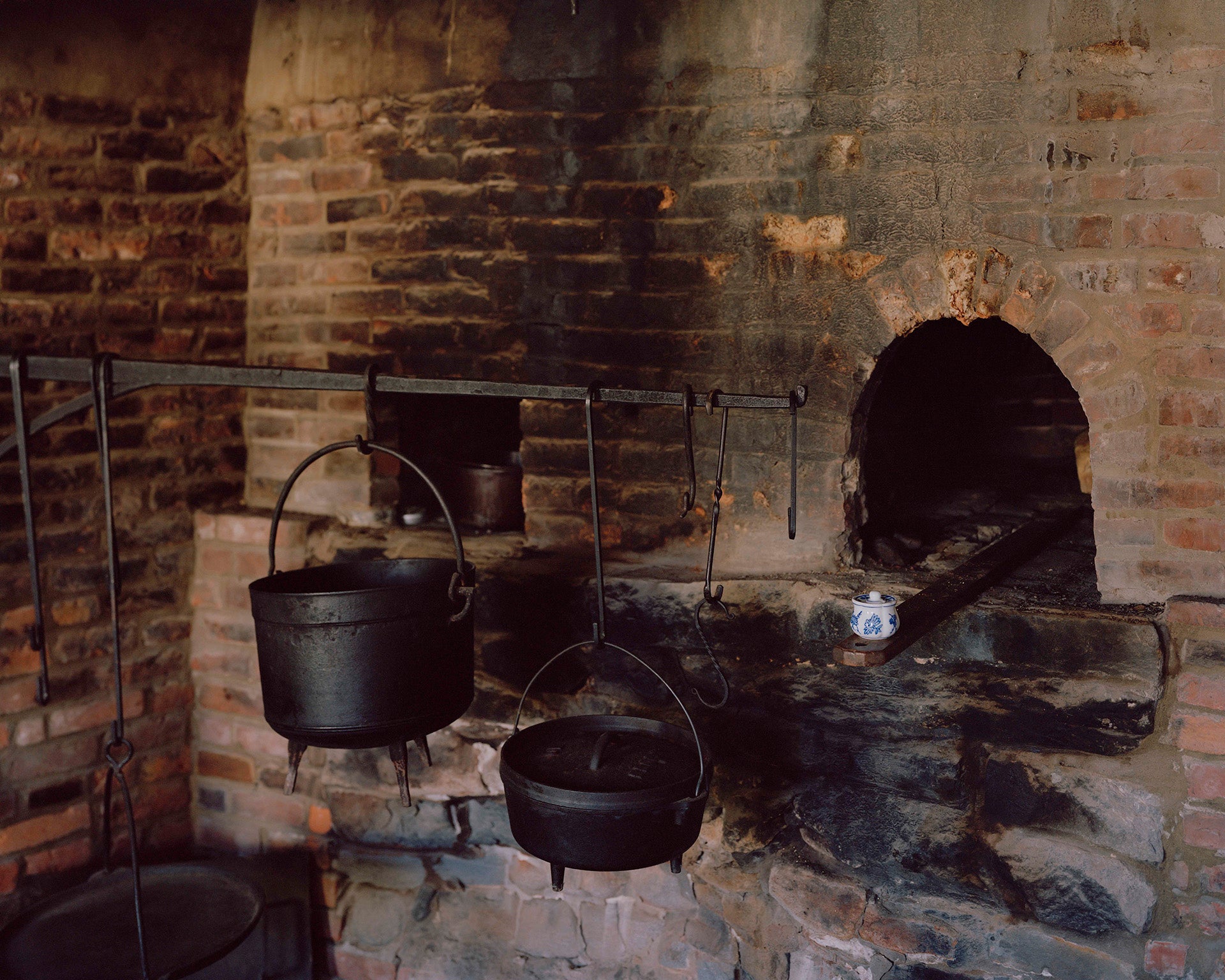 Photograph of brick oven hearth, with three cast iron pots hanging from a large rod. 