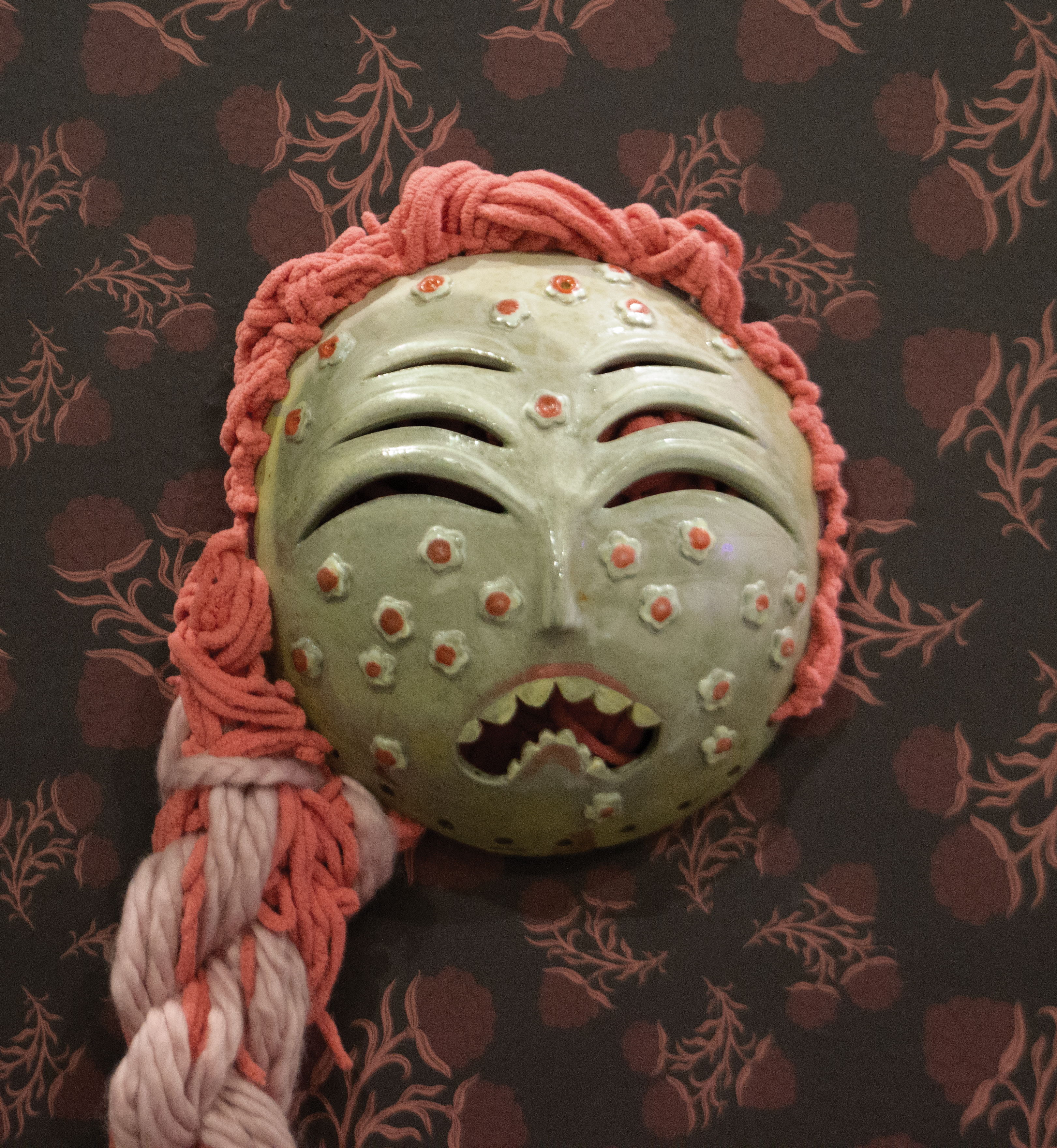 Green ceramic mask with six eyes, wailing mouth, and coral yarn hair.