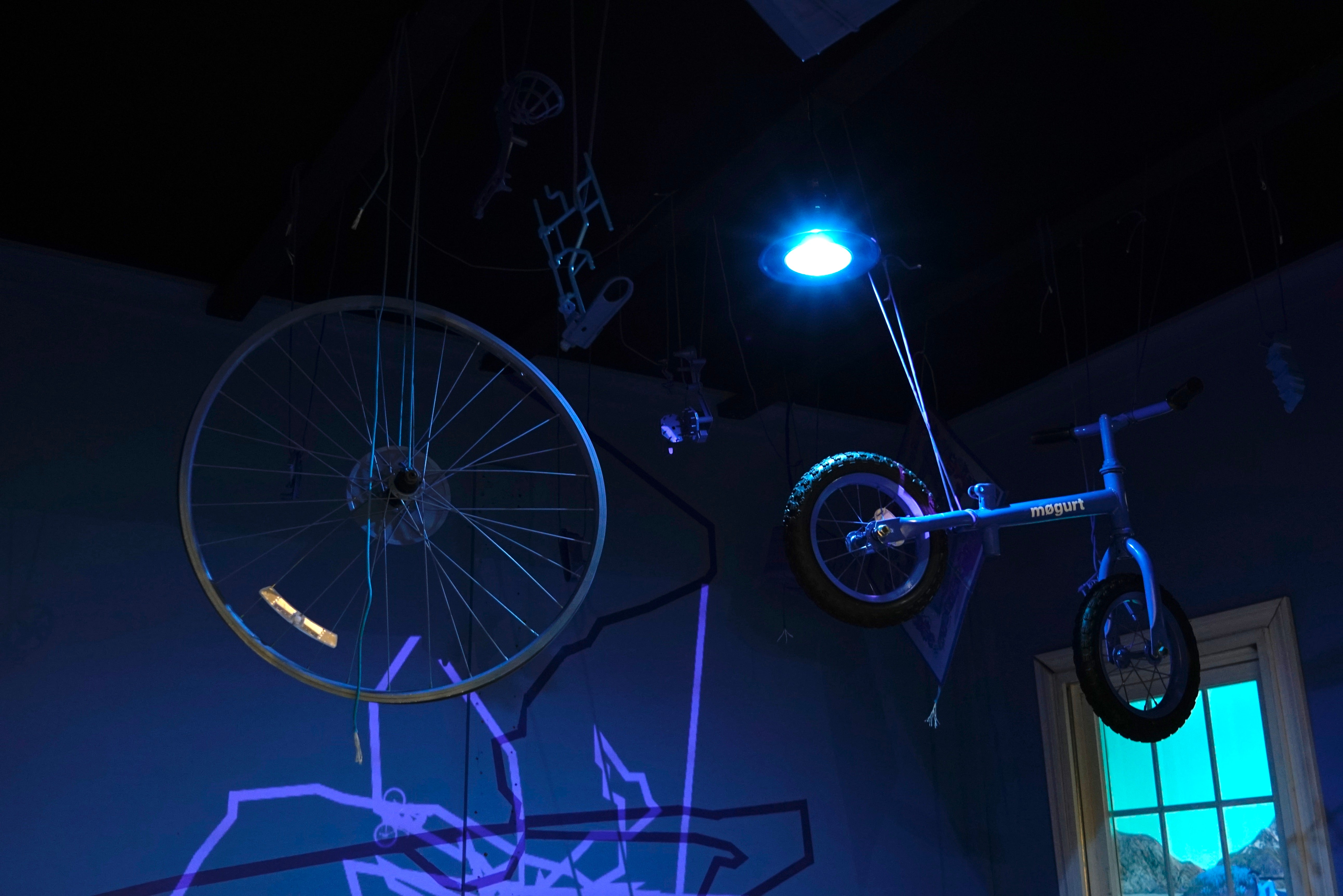 Installation materials suspended from the ceiling: Mouse trap board game pieces, kid's bike, busted bike wheel, paracord