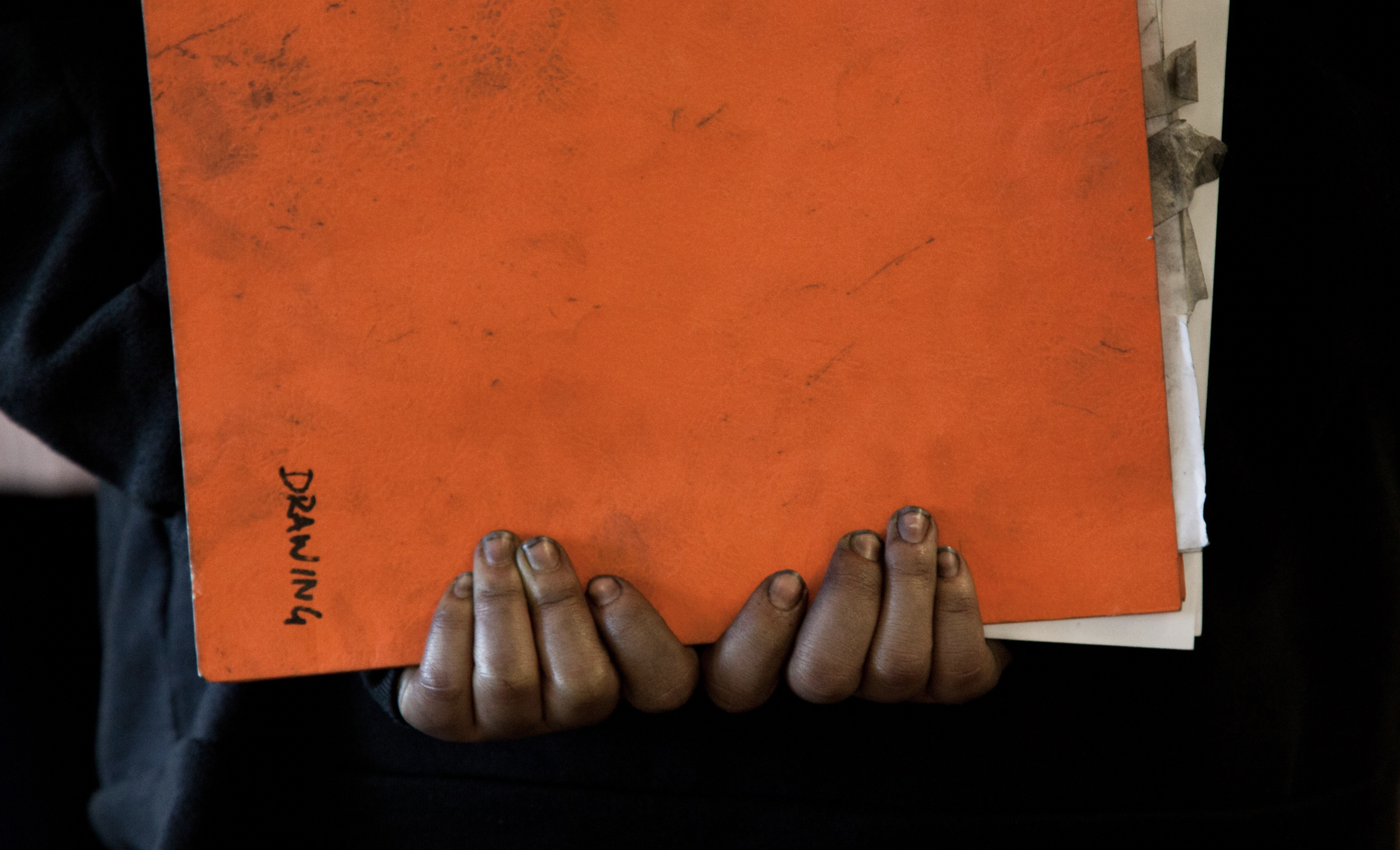 A pair of young Black hands grasp an orange folder labeled "Drawing"
