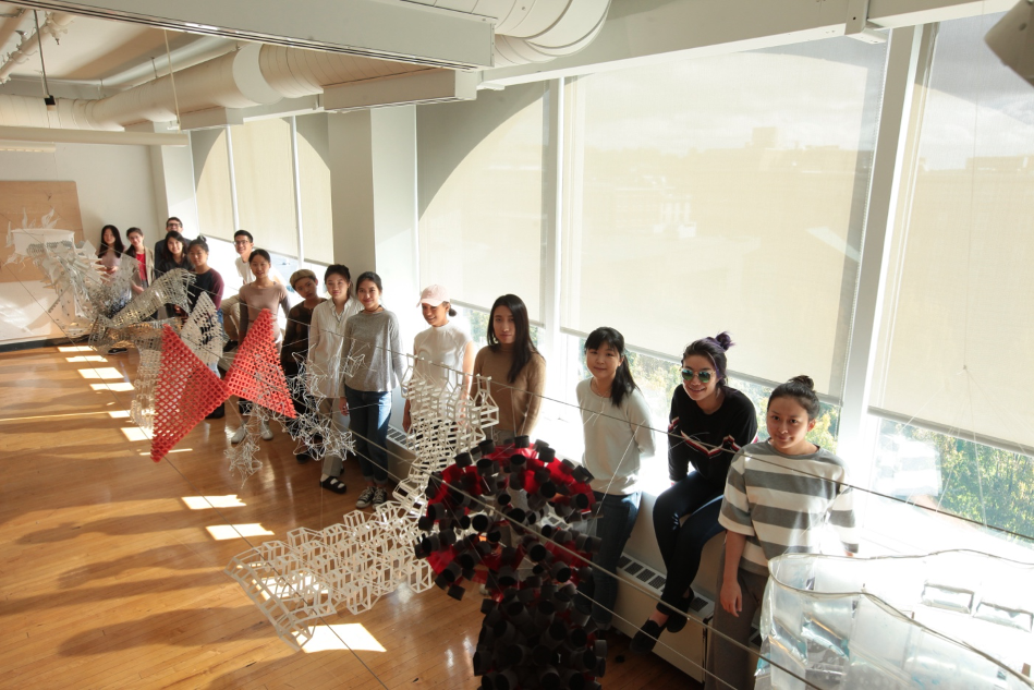 A row of students sit with 3D work in front of windows