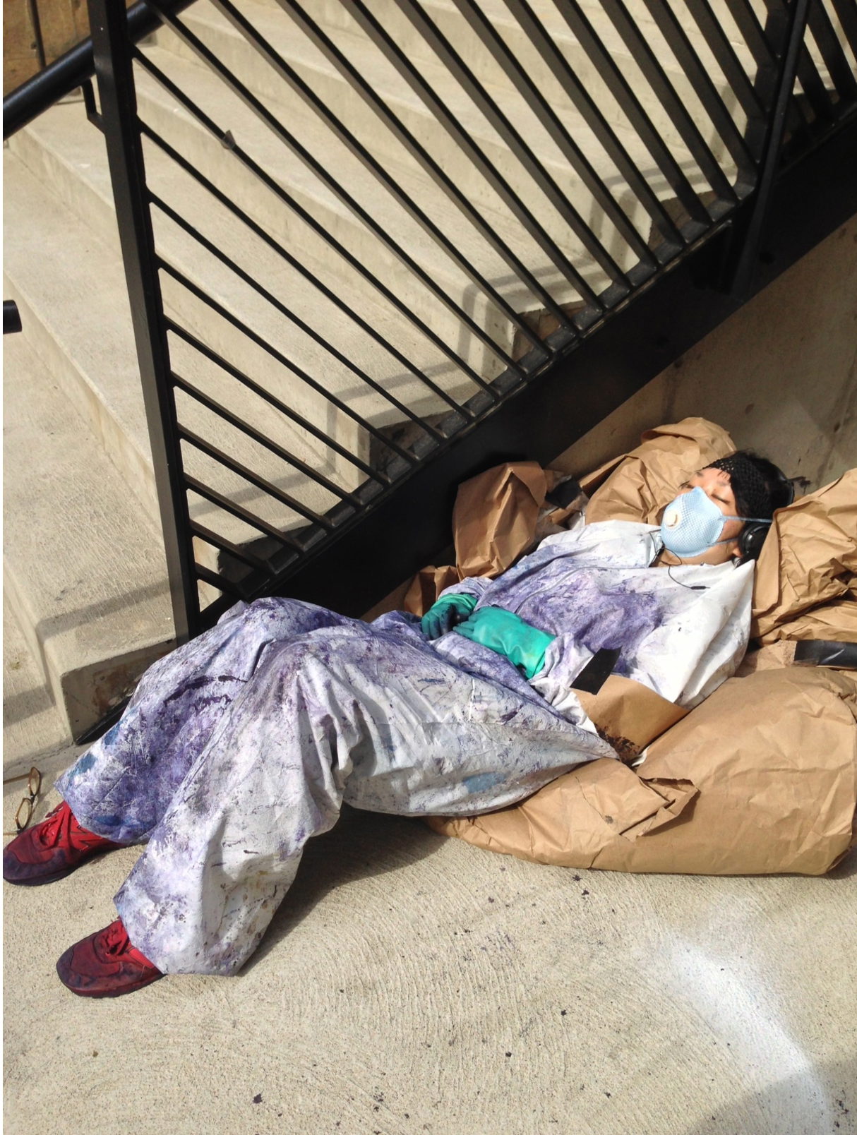 A tired student reclines on brown bags by stairs with a metal railing. They are wearing headphones, rubber gloves, a dye-splattered lab coat, and a respirator mask.