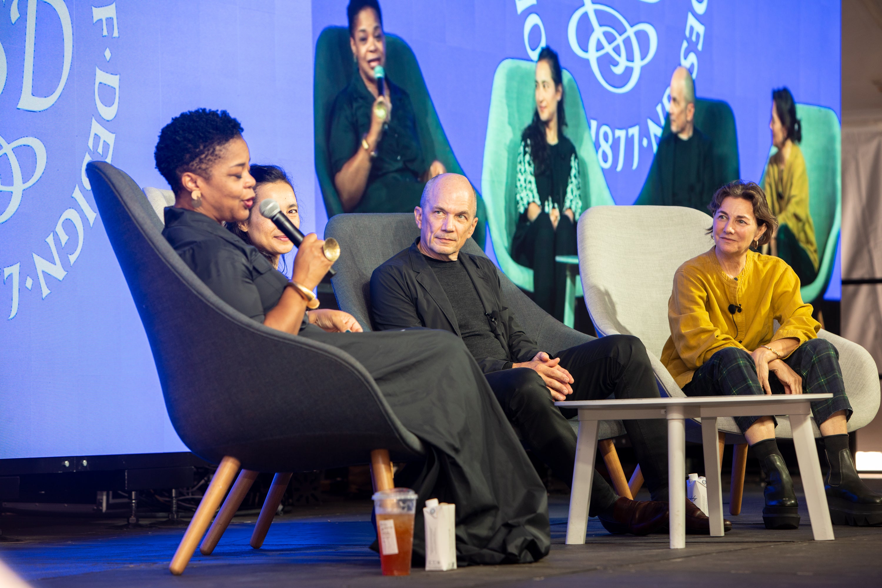 /Three%20women%20and%20one%20man%20sit%20in%20chairs%20on%20a%20stage.%20One%20of%20them%2C%20a%20Black%20woman%2C%20speaks%20into%20a%20microphone.%20They%20are%20projected%20onto%20a%20screen%20behind%20them.