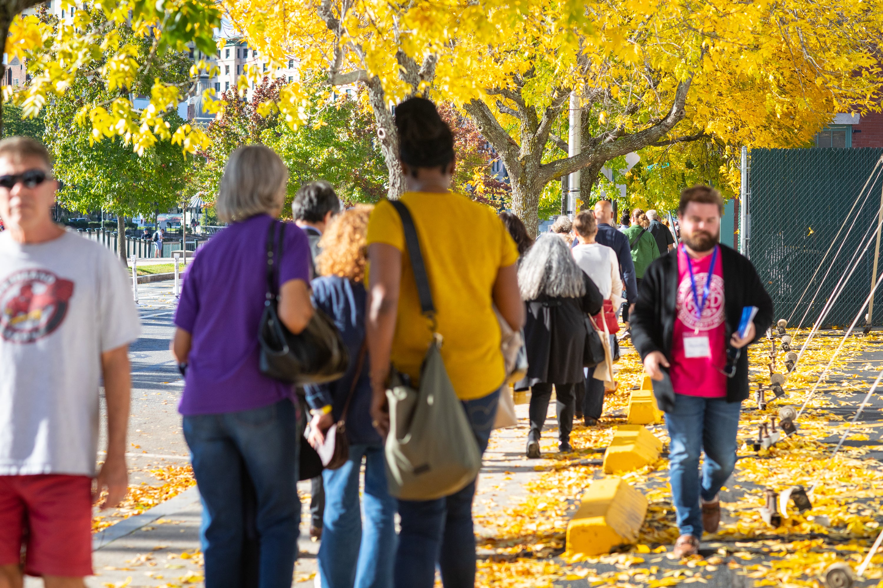 /Crowds%20of%20people%20walk%20down%20a%20tree-lined%20sidewalk.%20The%20trees%20are%20full%20of%20bright%20yellow%20leaves.