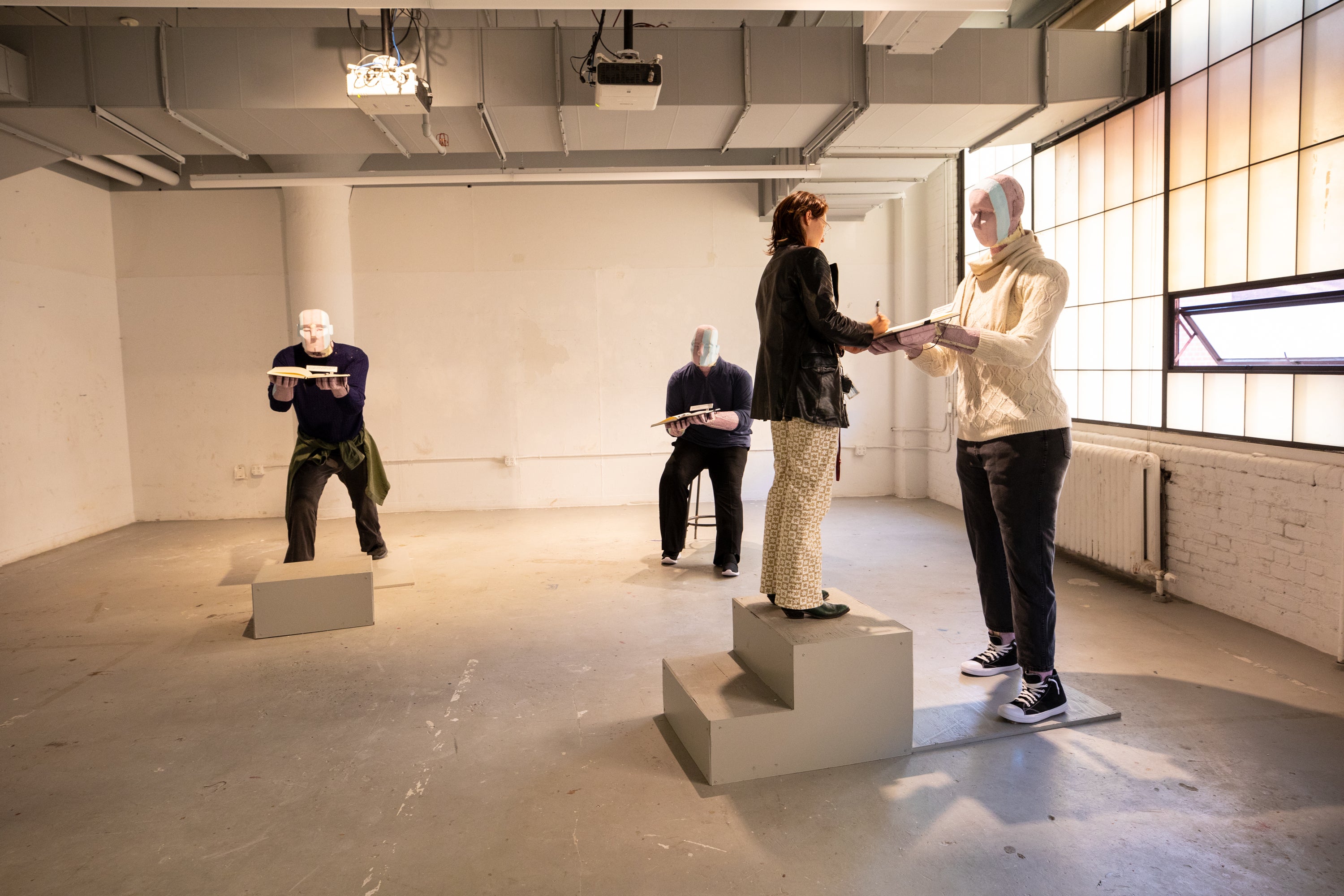 /White%20gallery%20space%20with%20three%20robotic-looking%20human-sized%20figures%20dressed%20in%20regular%20clothes%2C%20holding%20books.%20A%20real%20human%20woman%20writes%20in%20one%20of%20their%20books.%20