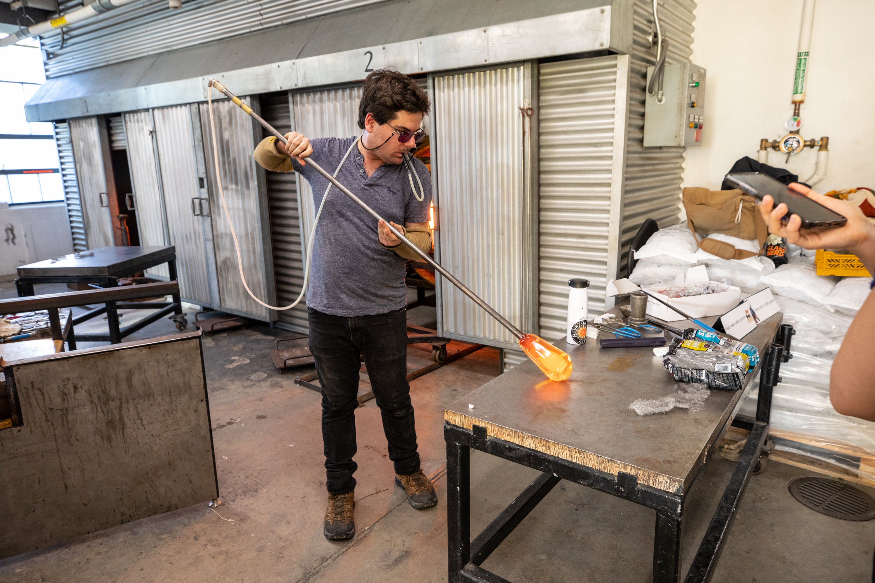 /A%20man%20demonstrates%20blowing%20glass.%20He%20holds%20a%20pipe%20with%20molten%20glass%20on%20the%20end%2C%20rolling%20it%20on%20a%20table%20as%20he%20blows%20into%20a%20tube%20connected%20to%20the%20pipe.