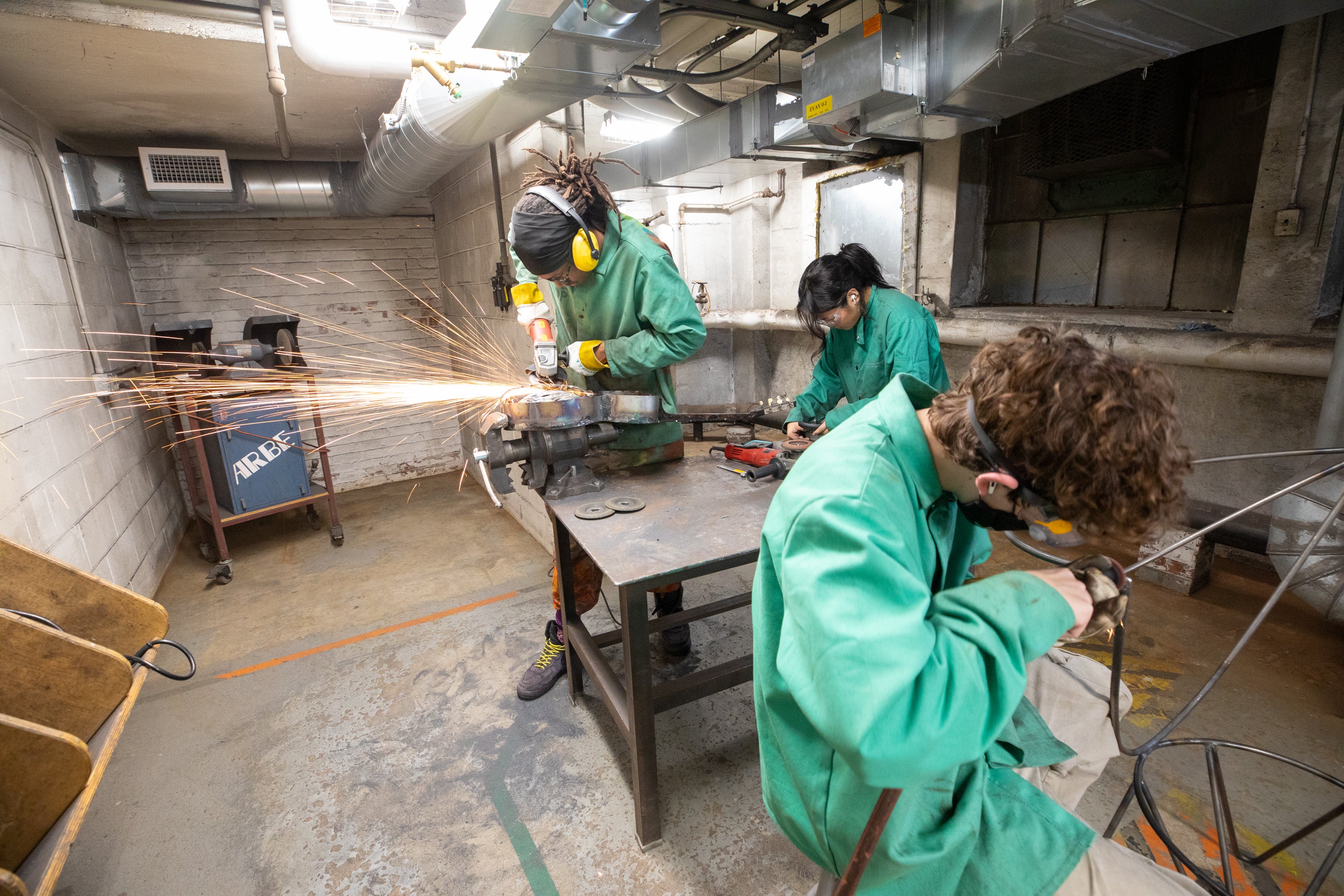 /Three%20students%20work%20in%20a%20metals%20shop%20wearing%20green%20protective%20smocks.%20Sparks%20fly%20as%20one%20student%20cuts%20through%20metal.