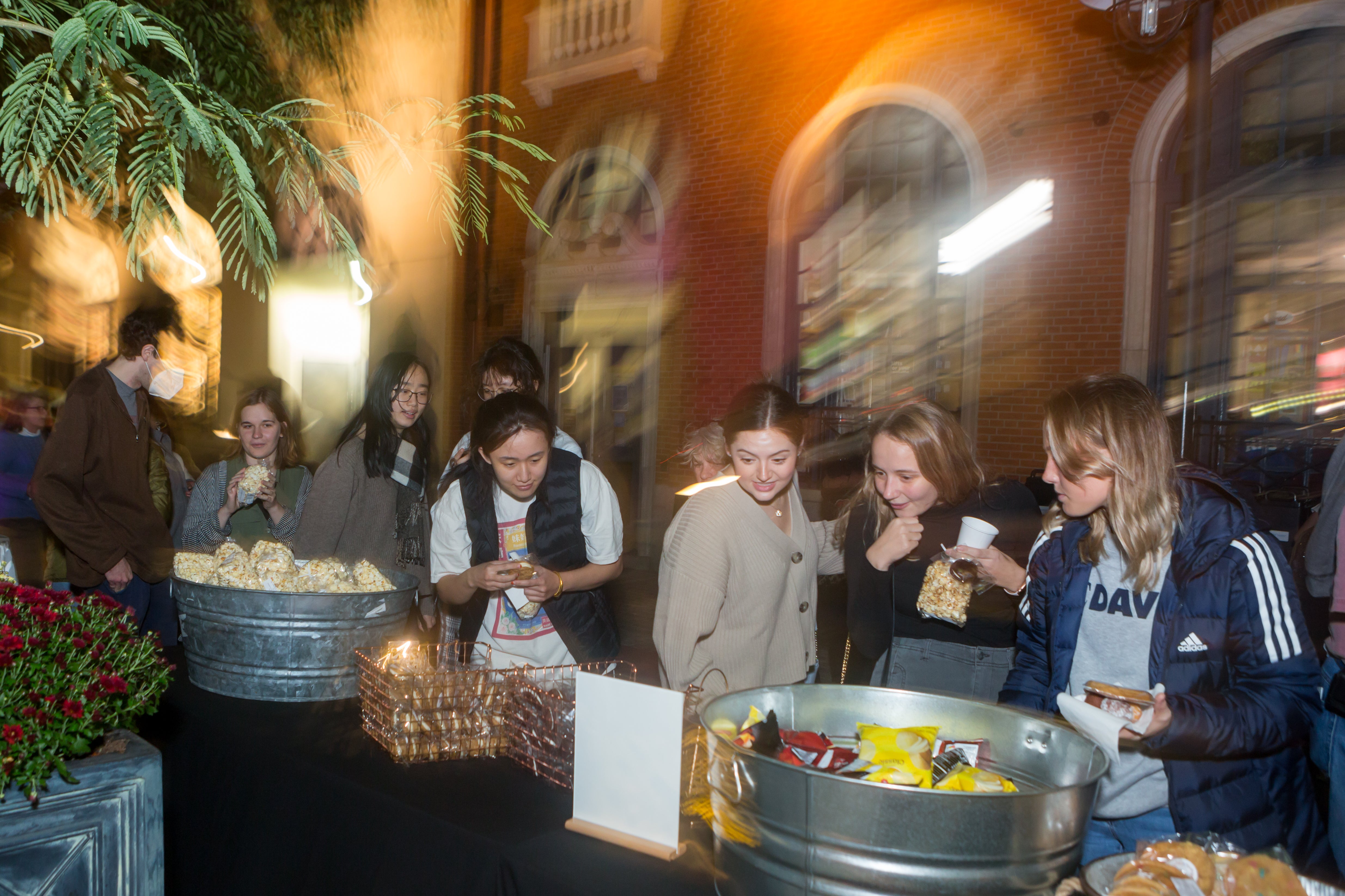 /Nighttime%20photo%20of%20several%20young%20women%20looking%20at%20a%20sign%20in%20front%20of%20a%20table%20of%20snacks.