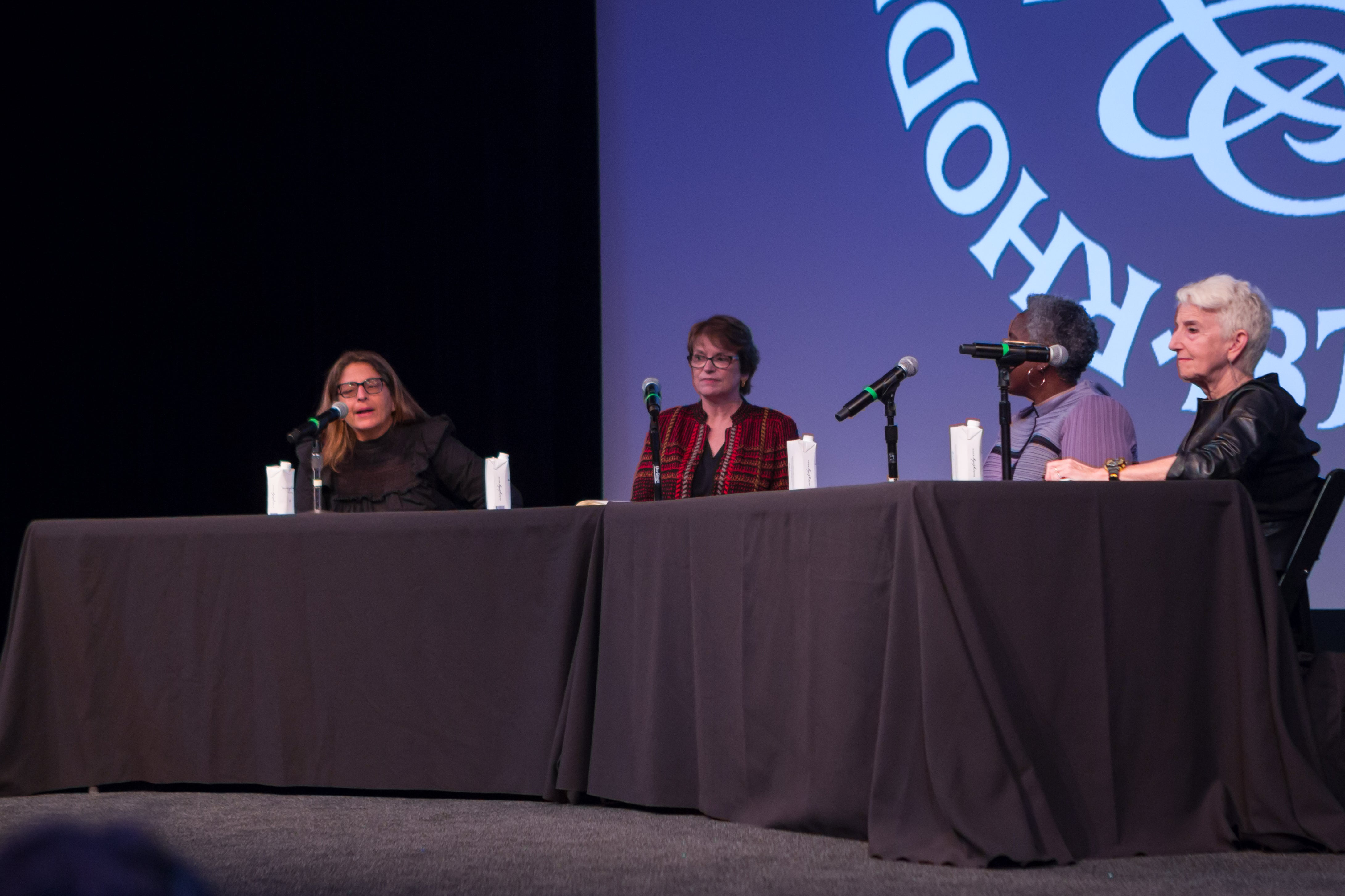 /Close-up%20shot%20of%20four%20women%20panelists%20on%20stage%20at%20a%20table%20draped%20in%20a%20black%20cloth.