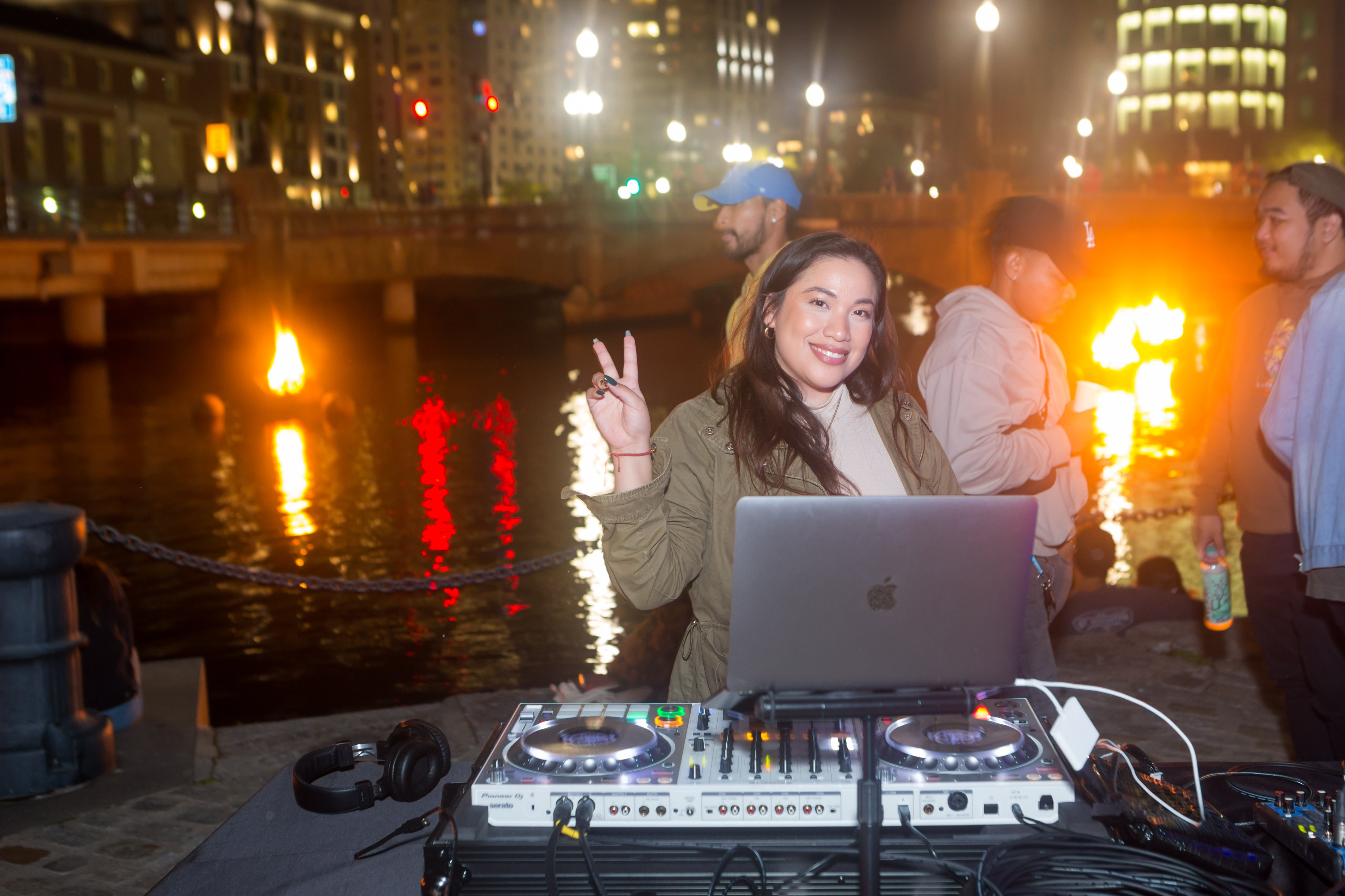 /At%20night%2C%20a%20smiling%20young%20woman%20stands%20in%20front%20of%20DJ%20equipment%2C%20raising%20her%20hand%20in%20a%20peace%20symbol.%20She%20has%20long%20green%20nails.%20In%20the%20background%2C%20braziers%20burn%20in%20the%20Providence%20river.