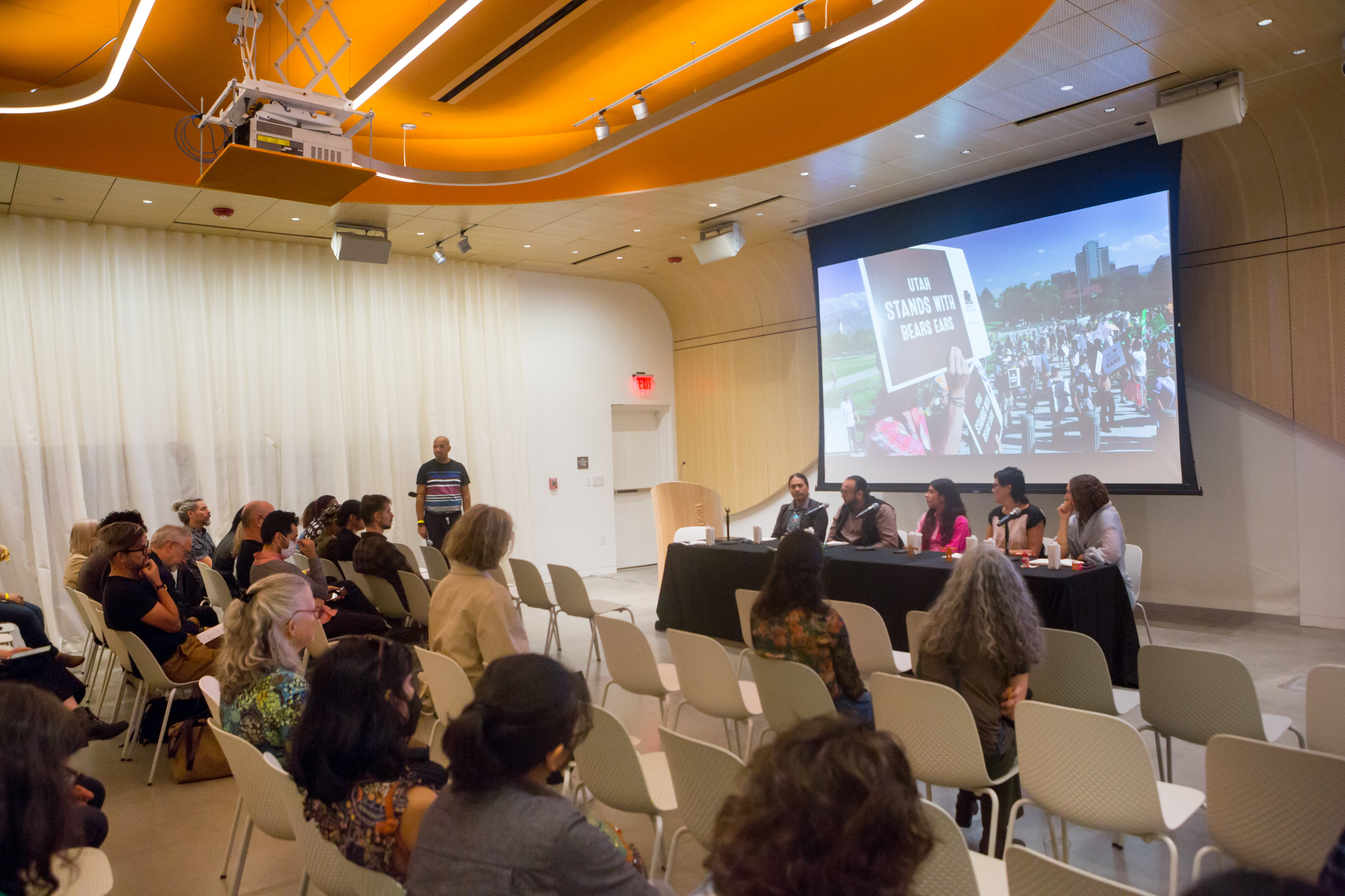 /A%20panel%20of%20five%20people%20present%20in%20front%20of%20a%20seated%20audience.%20A%20projection%20behind%20them%20shows%20a%20peaceful%20protest%20with%20someone%20holding%20a%20sign%20that%20says%20%27Utah%20stands%20with%20Bears%20Ears.%27