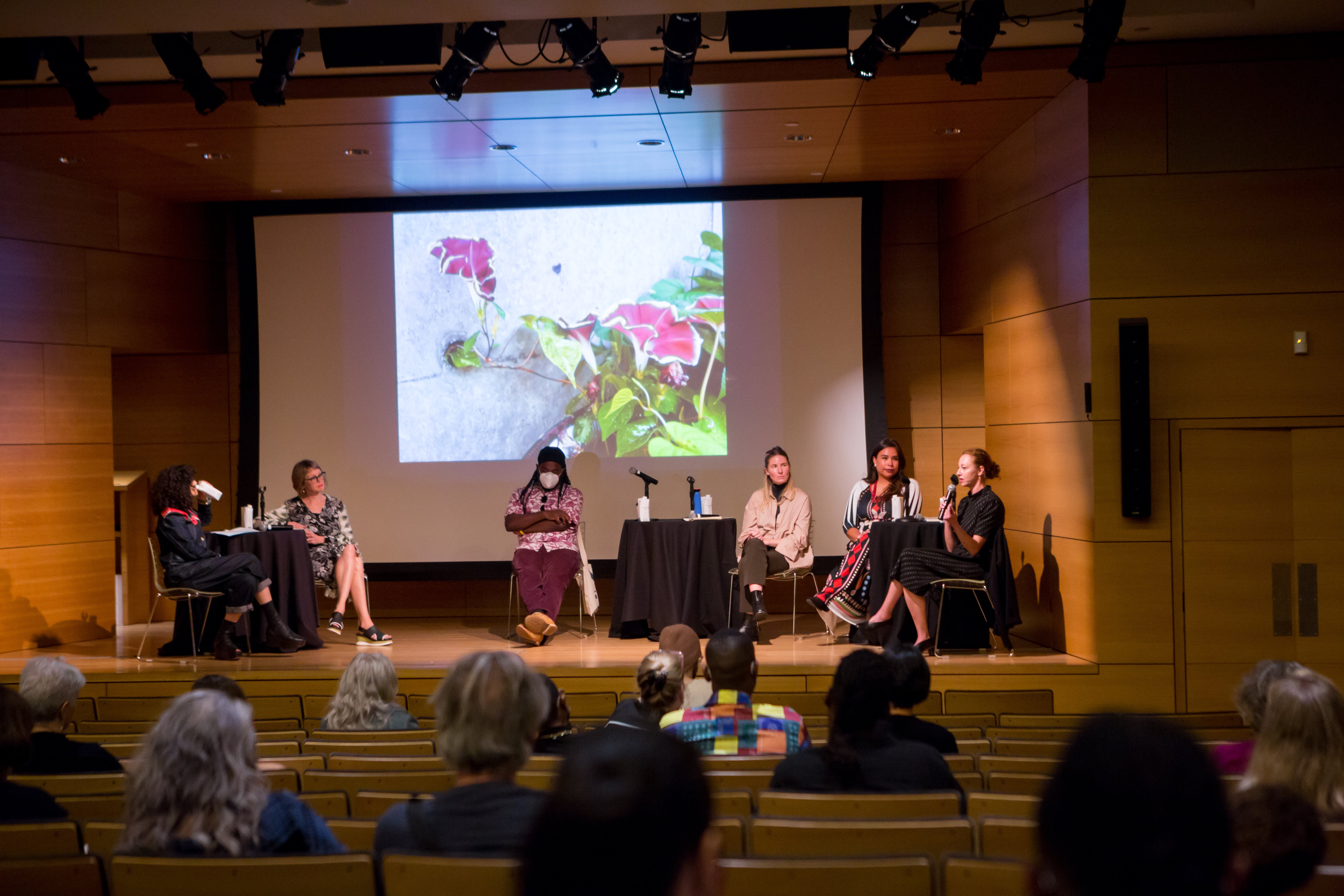 /Four%20panelists%20and%20two%20moderators%20sit%20on%20stage%20at%20a%20wood-paneled%20auditorium.%20An%20image%20of%20a%20flowering%20vine%20is%20projected%20behind%20them.